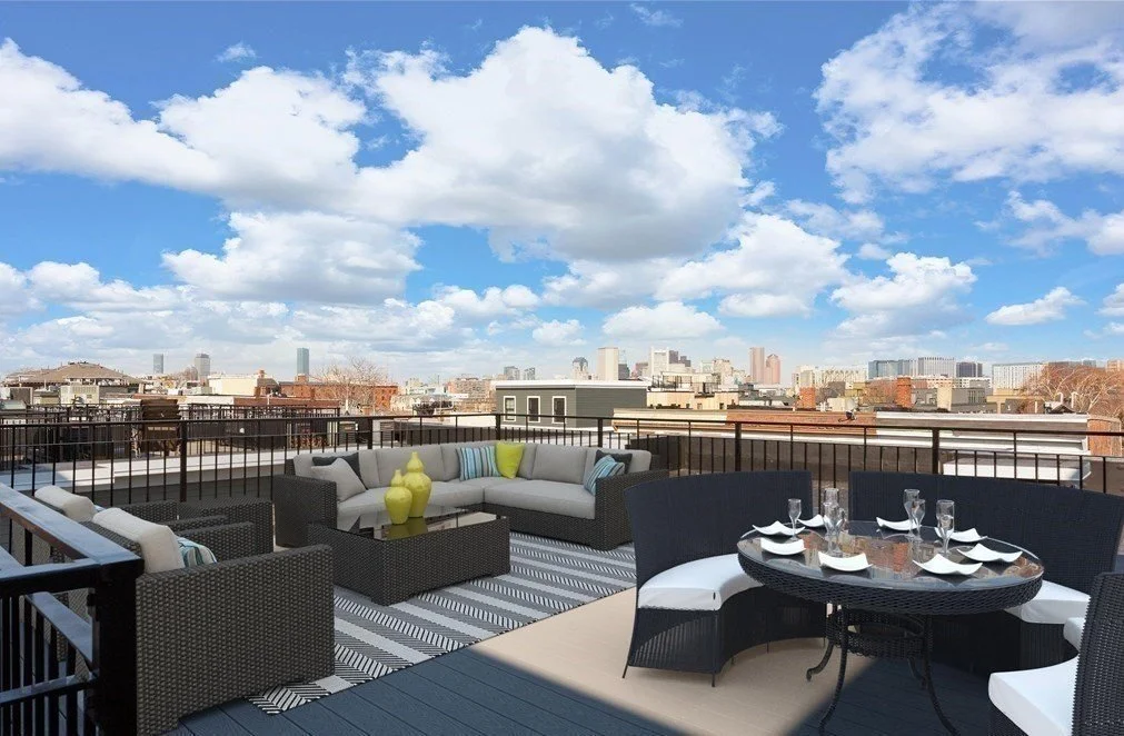 Rooftop terrace with outdoor wicker seating, dining table, and city skyline beneath a blue sky with clouds