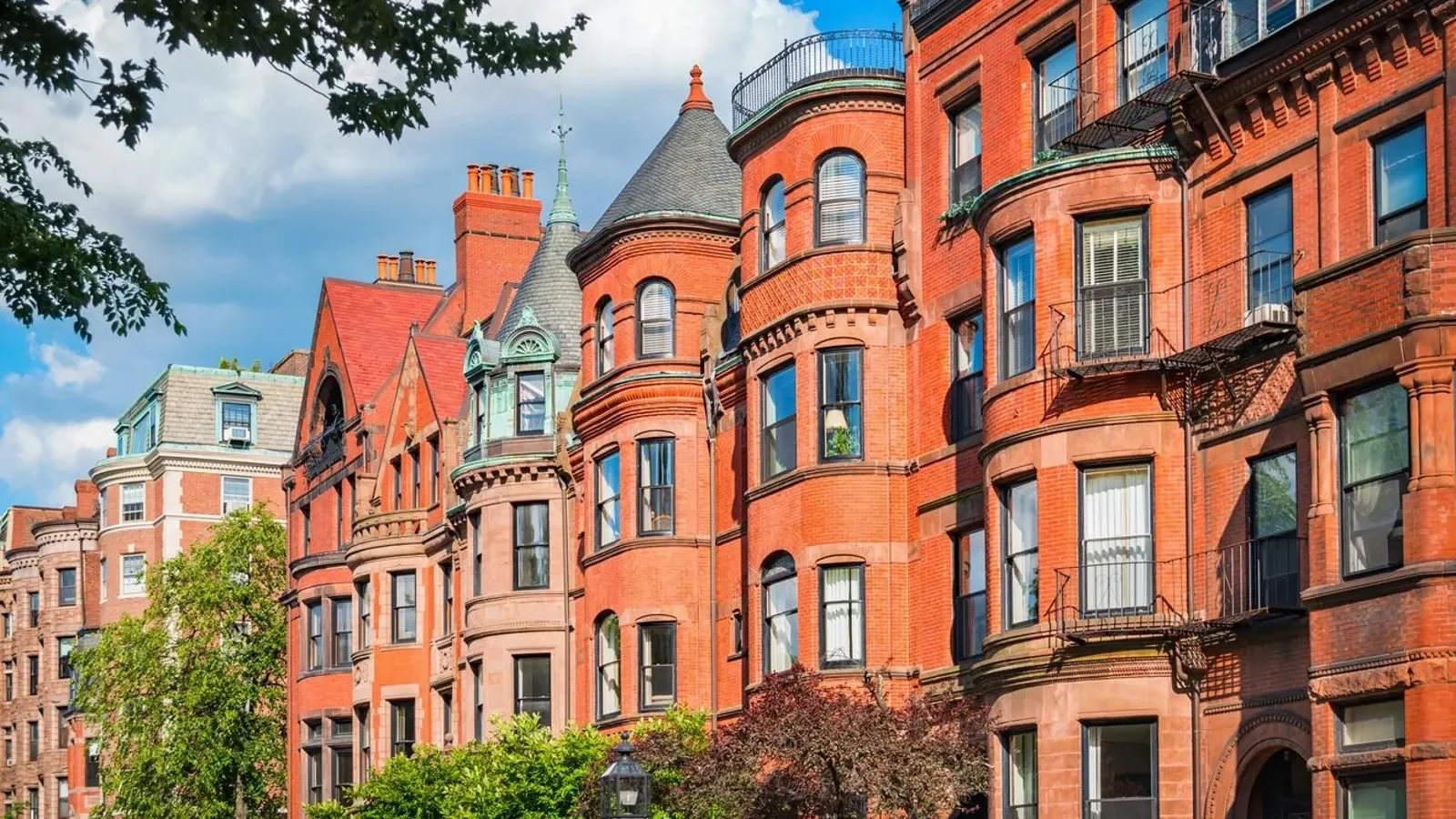 Row of ornate Victorian red-brick townhouses with bay windows, turrets, balconies, and chimneys under a blue sky