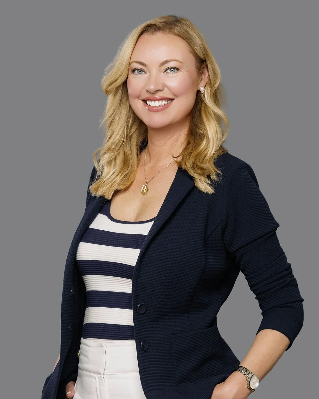 Smiling blonde woman in a navy blazer and striped shirt posing against a gray background