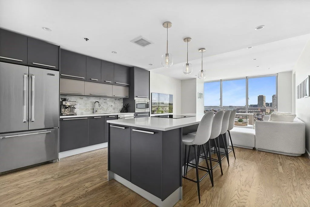 Modern condo kitchen with dark cabinets, stainless steel appliances, island seating, and city views through large windows