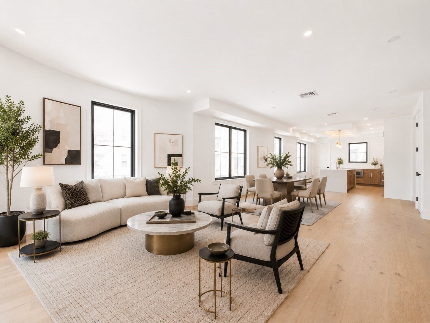 Bright open-concept living room and dining area with neutral furnishings, hardwood floors, large windows, and adjoining kitchen