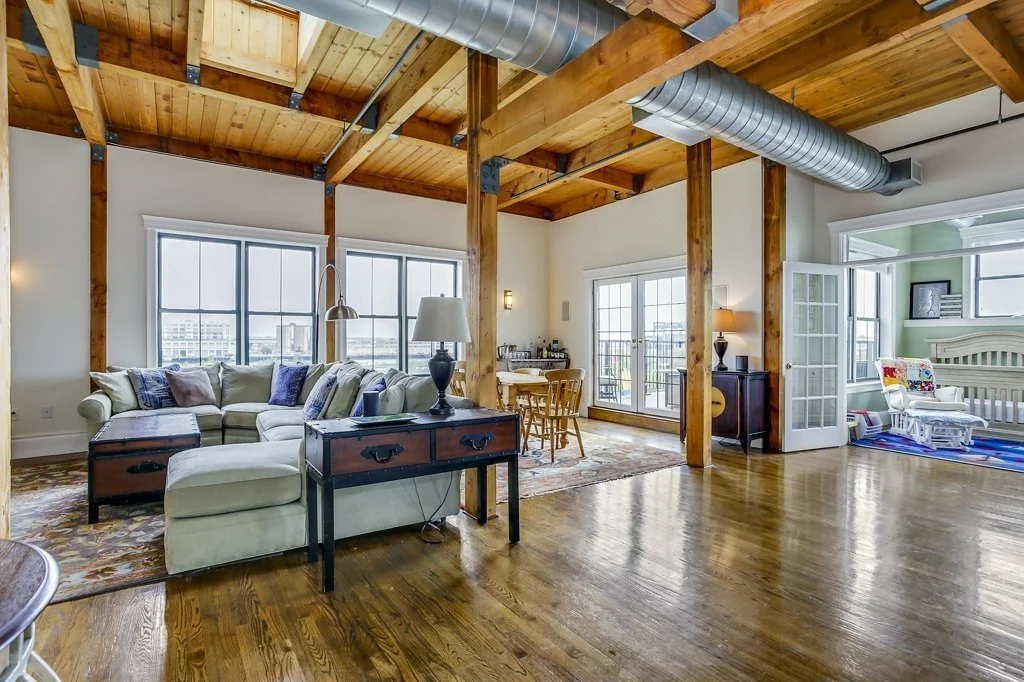 Open-concept industrial loft living room with exposed wood beams, polished hardwood floors, sectional sofa, dining area, and exposed ductwork