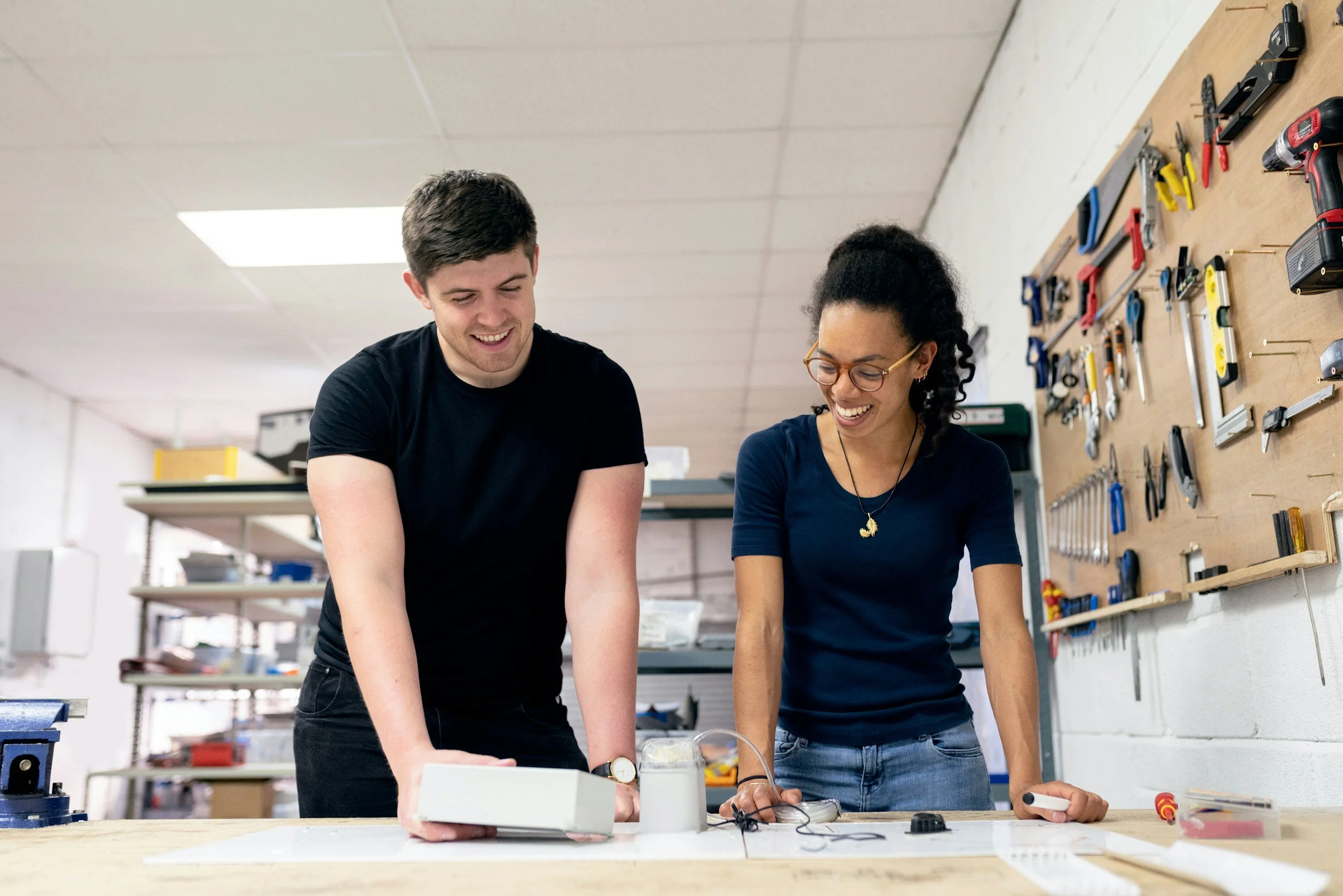 Two people working together in a workshop, smiling and looking down at a work table with tools and electronic equipment, with a pegboard full of tools on the wall behind them.