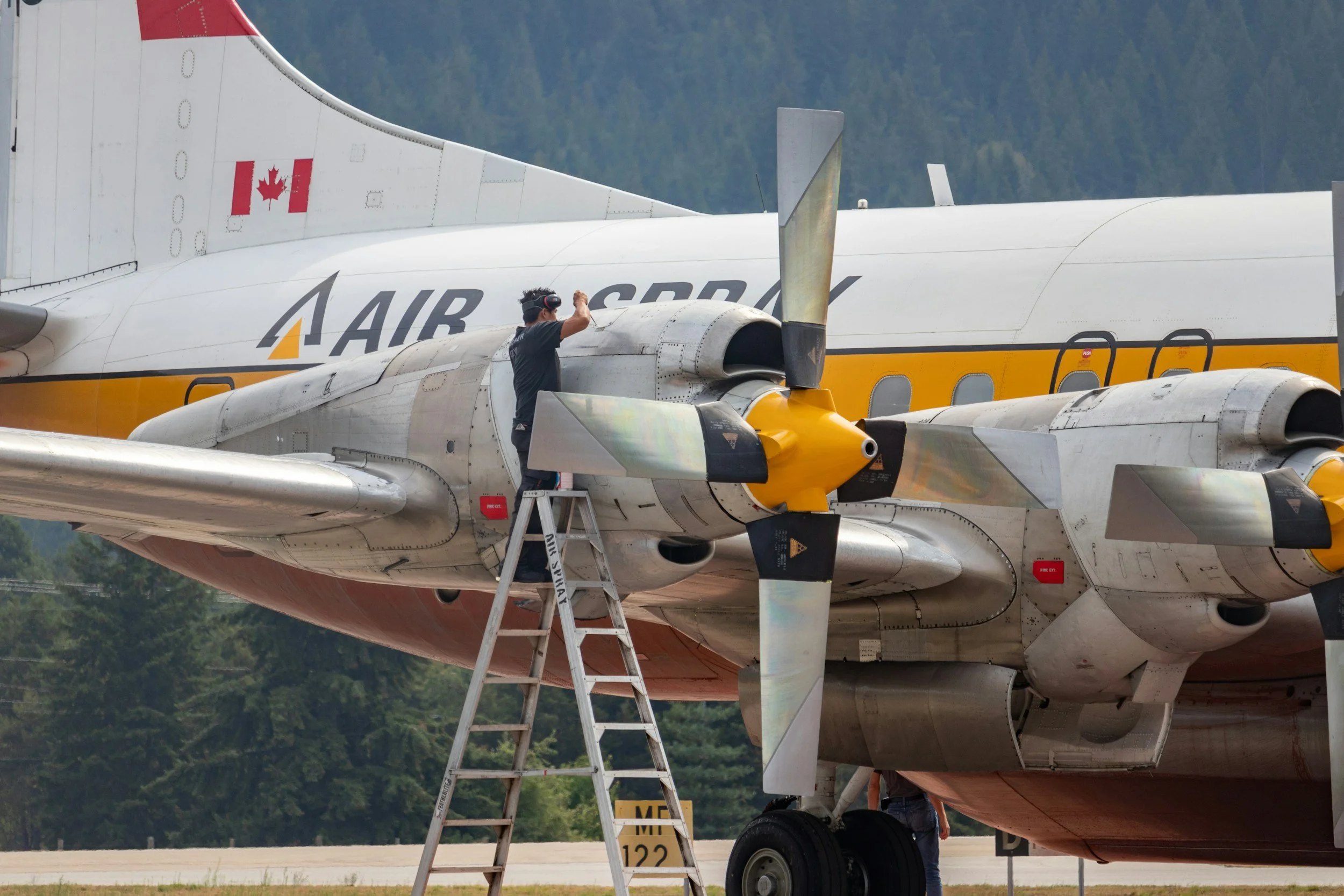 A person wearing a headset standing on a ladder inspecting or working on the engine of a large military aircraft with the word 'AIR' visible on the fuselage, and a Canadian flag on the tail fin, outdoors with trees in the background.
