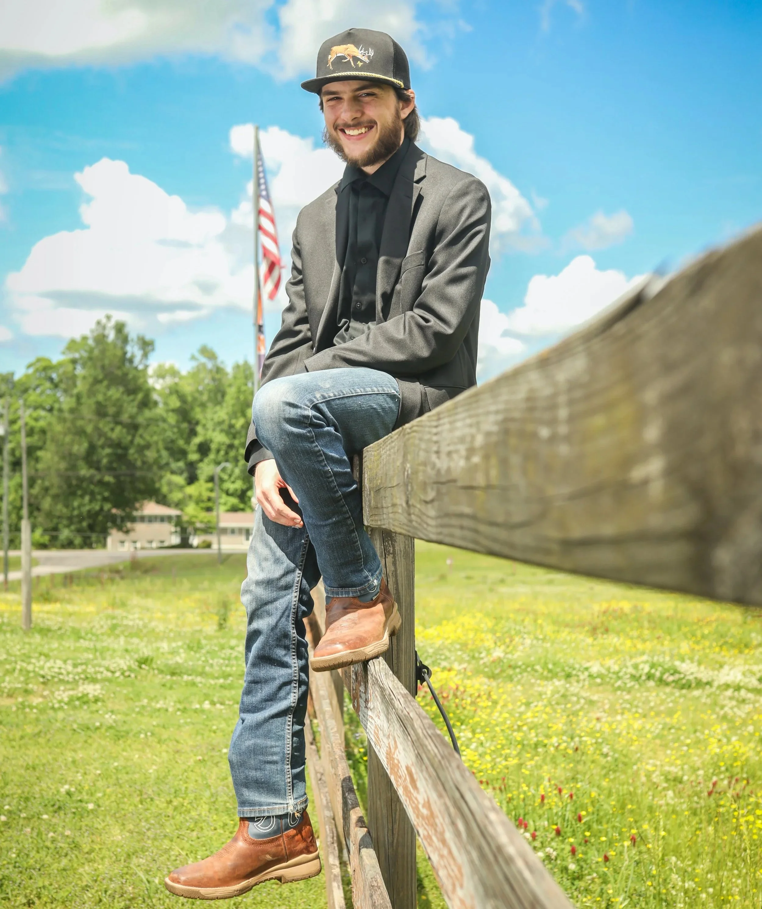 Clayton Mark, a young man with a beard and long hair, smiling in a suit jacket, black shirt, jeans, and brown boots, sitting on a wooden fence in a park with trees and yellow flowers, with a partly cloudy sky and an American flag in the background.