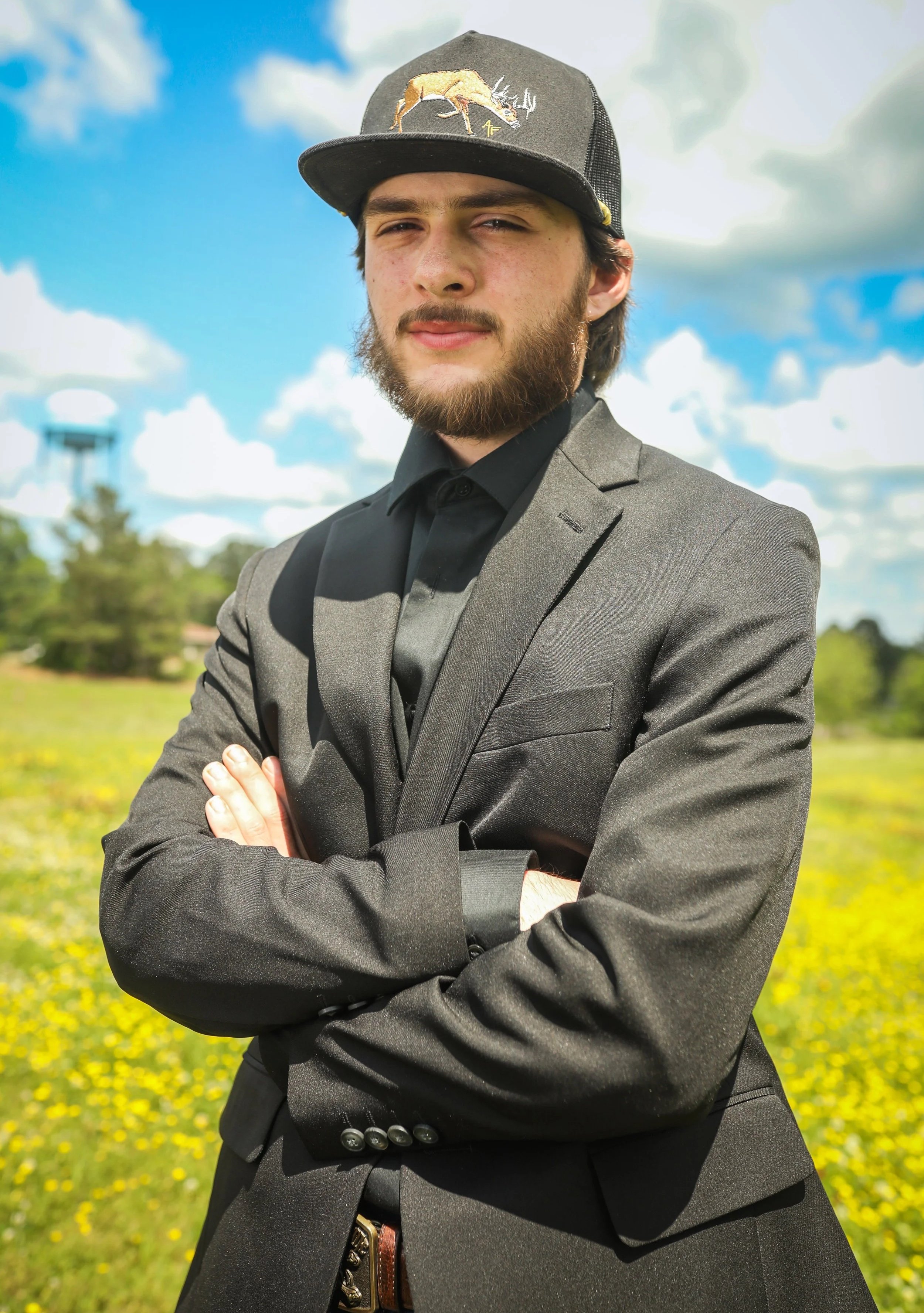Clayton Mark, a young man wearing a black suit, black shirt, and a black baseball cap, standing outdoors in a grassy area with yellow flowers, trees, and a partly cloudy sky in the background.