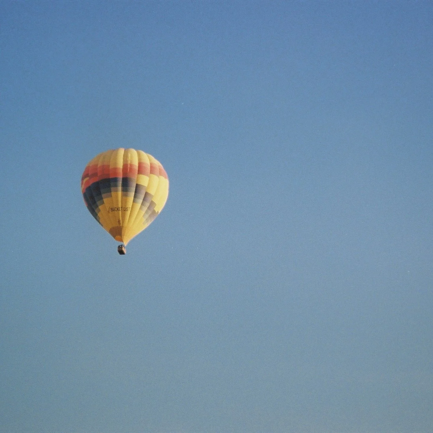 A hot air balloon with a yellow, red, black, and gray pattern floating in a clear blue sky.