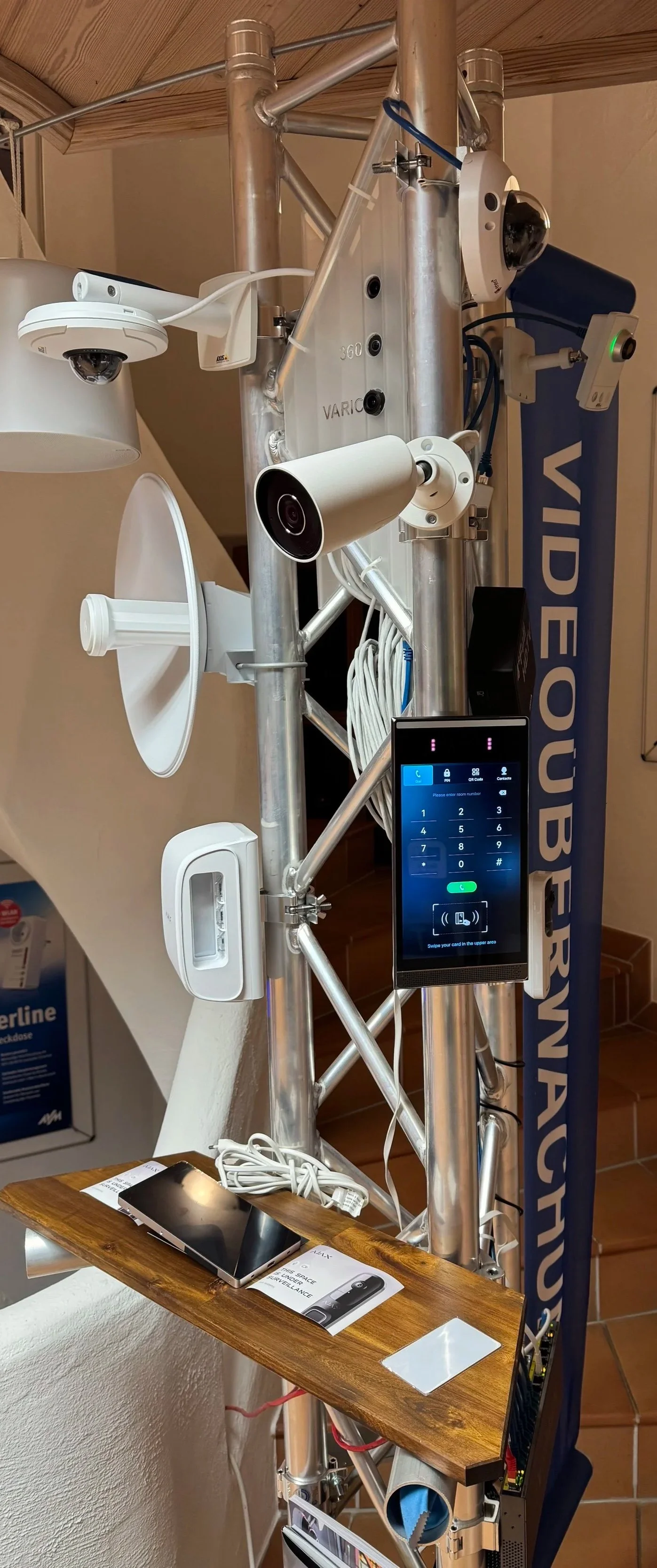 Technisches Gerät mit verschiedenen Kameras, einem Bildschirm und Kabeln, montiert auf einem Metallrahmen, daneben eine Holzablage mit einem Smartphone, Kabeln und Papieren.