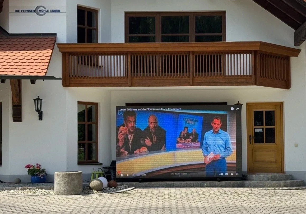 Ein Haus mit einem Balkon und mehreren Fenstern. Vor dem Haus steht ein Fernseher, auf dem eine Sendung mit mehreren Personen zu sehen ist, und eine Person, die vor dem Bildschirm steht.