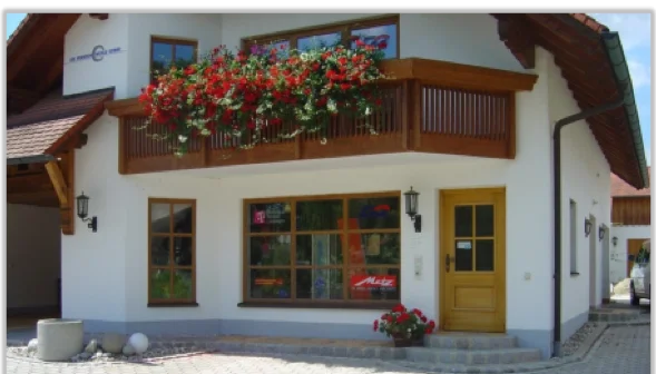 Ein zweistöckiges Haus mit weißer Fassade, Holzbalkon mit Blumen und gelber Tür im Vordergrund.