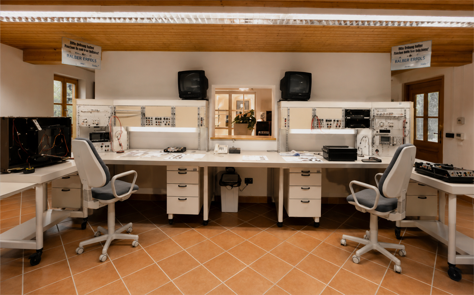 Ein Technikerarbeitsplatz mit Computern, Werkzeugen und elektrischen Komponenten in einem Raum mit weißen Wänden, Holzbalkendecke und Fliesenboden, die Fenster lassen natürliches Licht herein.