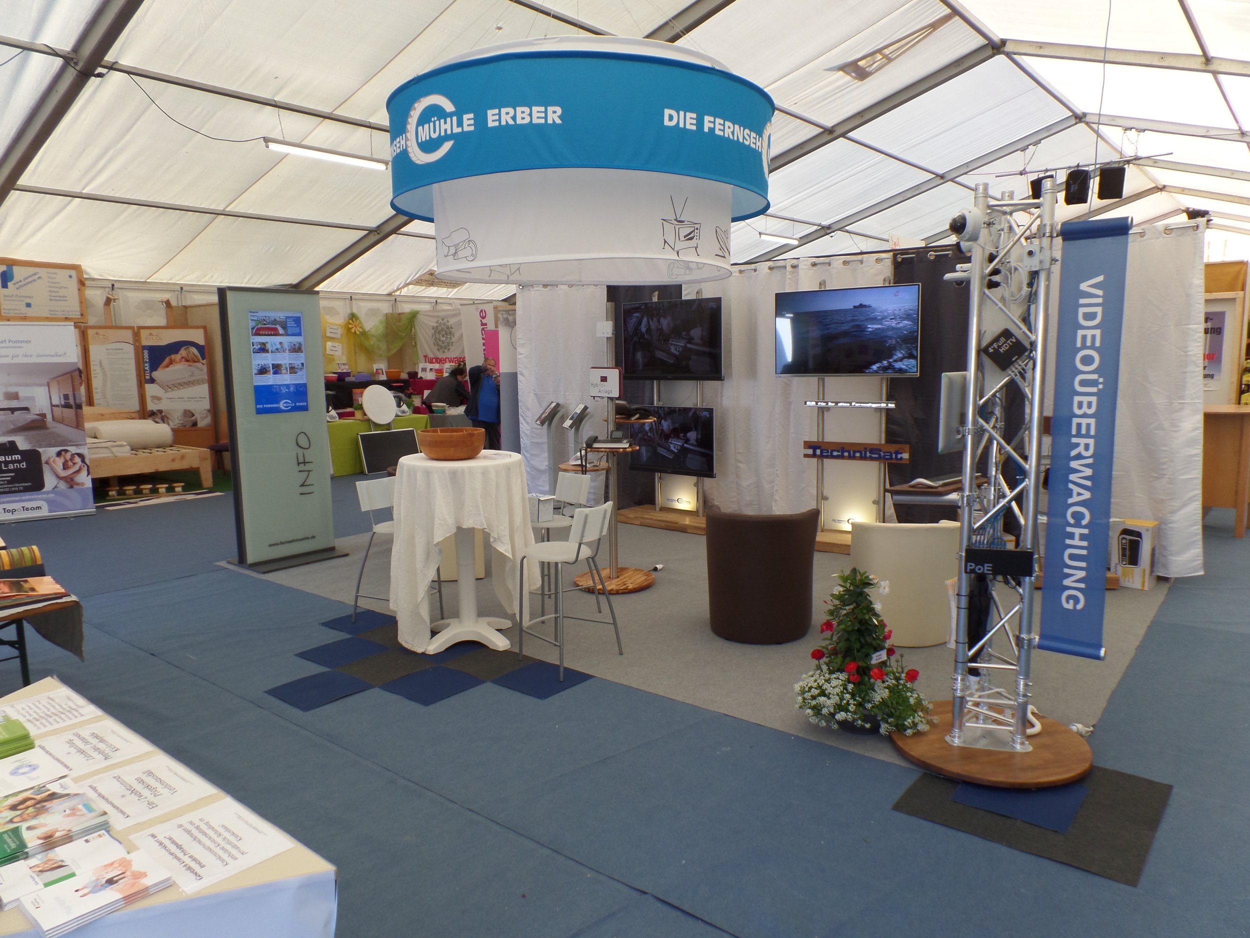 Innenaufnahme einer Messehalle mit einem Stand, der Geräte und Bildschirme zeigt. Es gibt einen runden Deckenhänger mit blauer Umrandung und weißem Text. Im Hintergrund sind mehrere Stände und Besucher sichtbar.