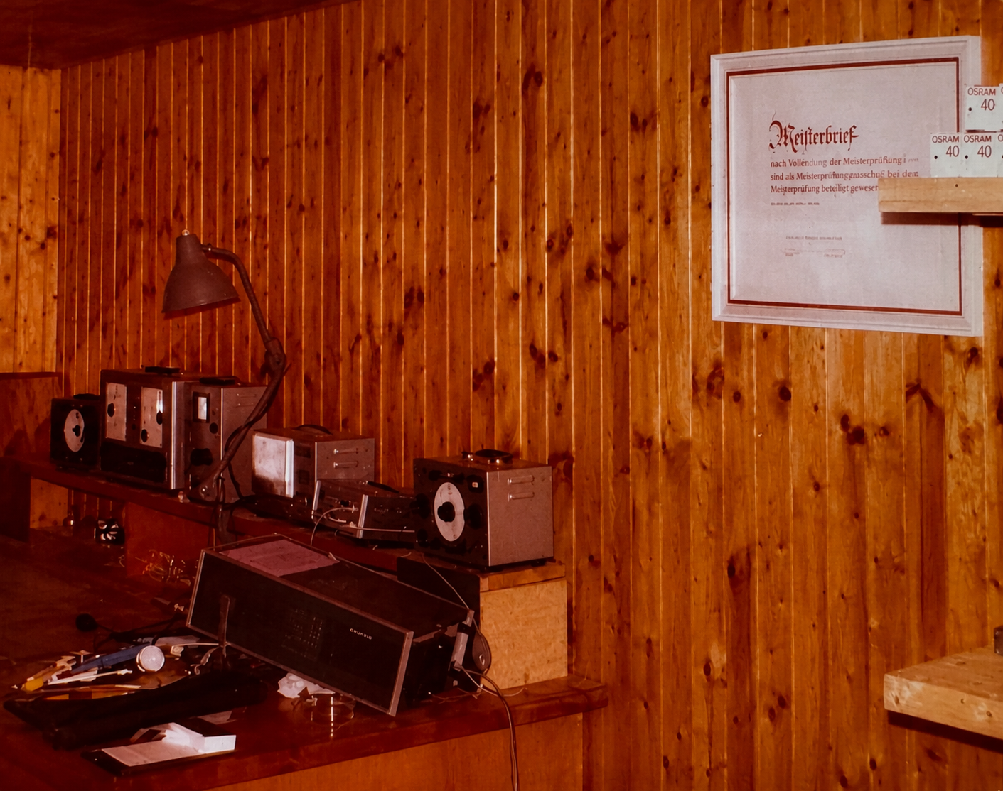 Ein Raum mit einer Holzwand und einem alten radioartigen Tonbandgerät auf einem Tisch. Es liegen Papier, Kabel und ein Mikrofon auf dem Tisch.