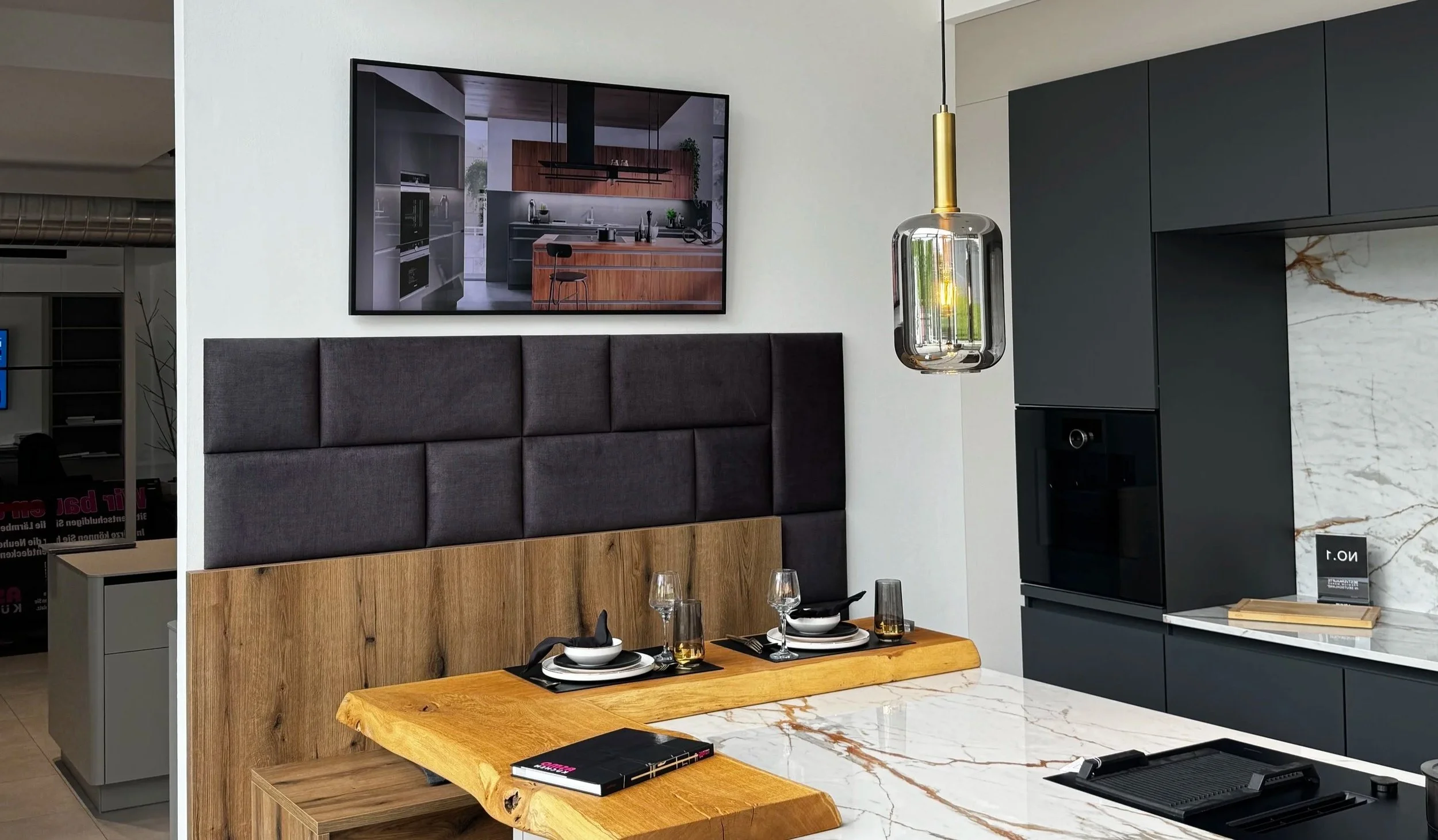 Moderne Küchen- und Essbereich mit dunklen Schränken, Holztisch, gestaffelte Beleuchtung und Fernseher an der Wand.