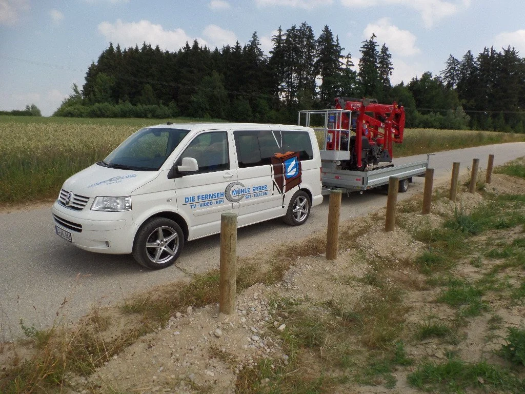 Weißer VW-Bus mit Werbung, der an einer Straße parkt, hinter dem ein Anhänger mit einem roten Feuerwehrauto steht; umgeben von Feldern und Bäumen im Hintergrund.