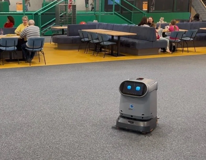 Ein Service-Roboter in einer modernen Lounge, im Hintergrund Sitzgelegenheiten mit mehreren Personen, grauer Teppichboden, farbige Wände und Treppen.