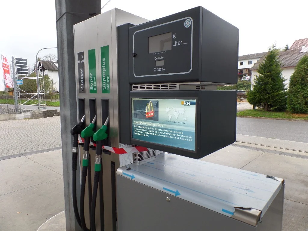 Tankstelle mit drei Zapfsäulen für Diesel, Super und Super Plus, mit elektronischer Anzeige und digitaler Bezahlbox, draußen vor einer Straße mit Häusern und Bäumen.