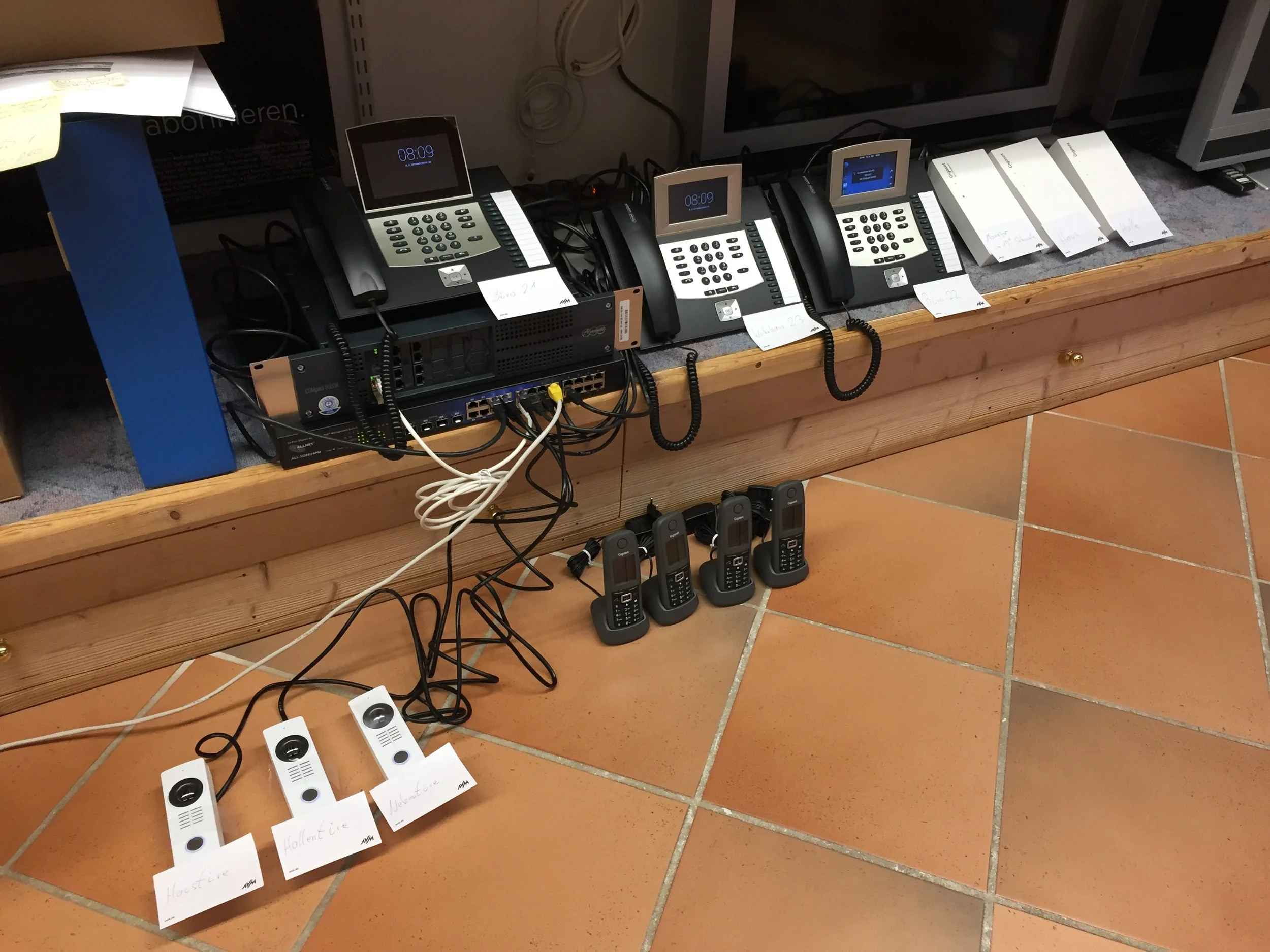 Mehrere schwarze und weiße alte Telefonmodelle mit Kabeln stehen auf einem Holzregal. Darunter sind auf Papierscheiben die Wörter 'Hollentic' und 'Hausatic' geschrieben. Es gibt auch drahtlose Mobiltelefone auf einem Tisch.