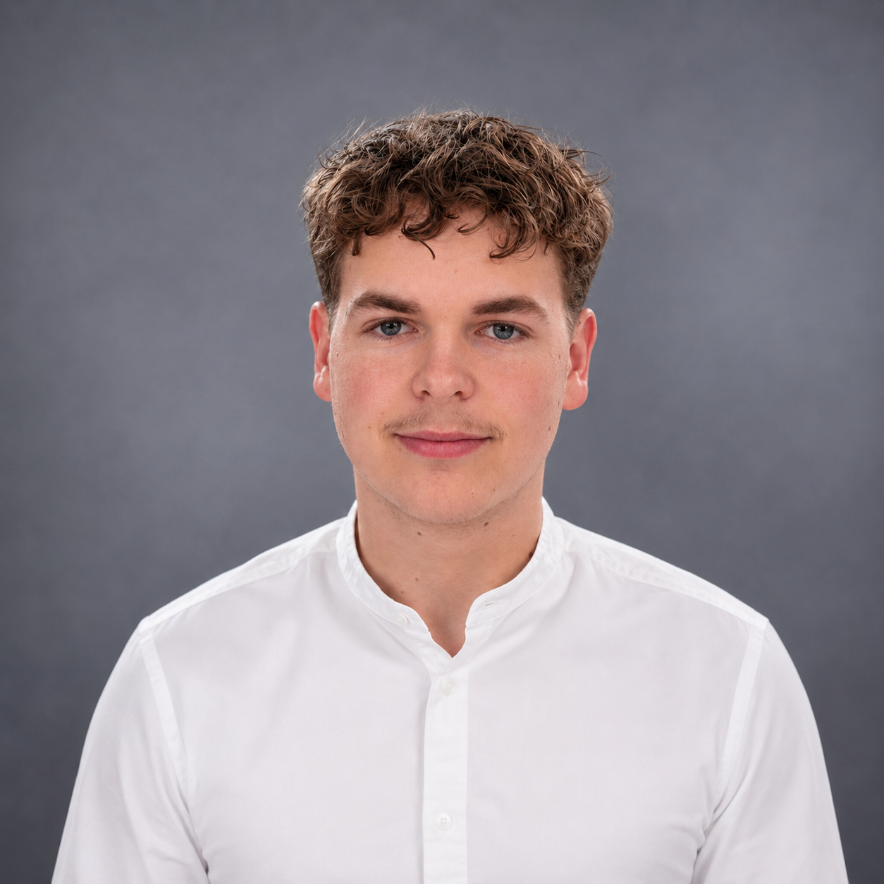 Porträt eines jungen Mannes mit braunen, gewellten Haaren, blauen Augen und einem leichten Lächeln, trägt ein weißes Hemd vor grauem Hintergrund.