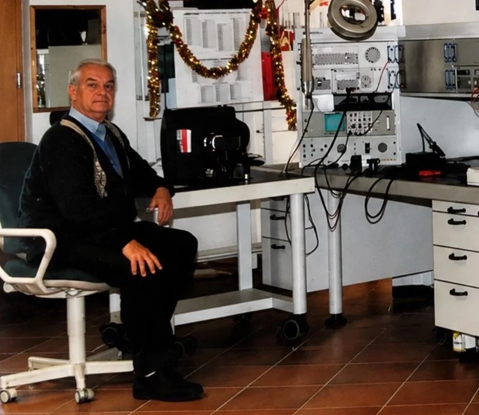 Ein Mann sitzt in einem Labor oder Werkstatt, umgeben von elektronischen Geräten und Kabeln, mit weihnachtlicher Dekoration im Hintergrund.