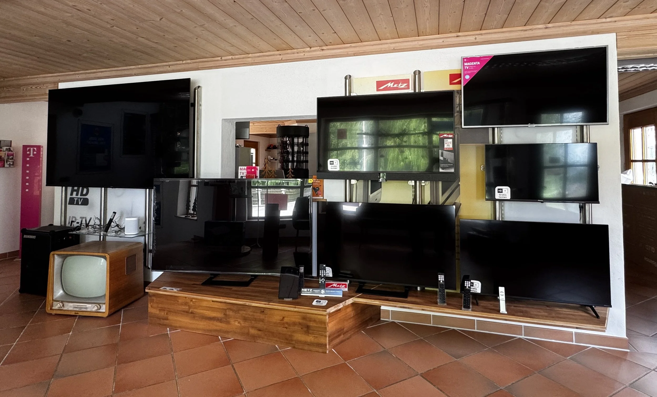 Mehrere Fernseher verschiedener Größen und Marken in einem Elektronikgeschäft auf einem Holzständer und Display-Regalen, mit Preisschildern und Produktinformationen.