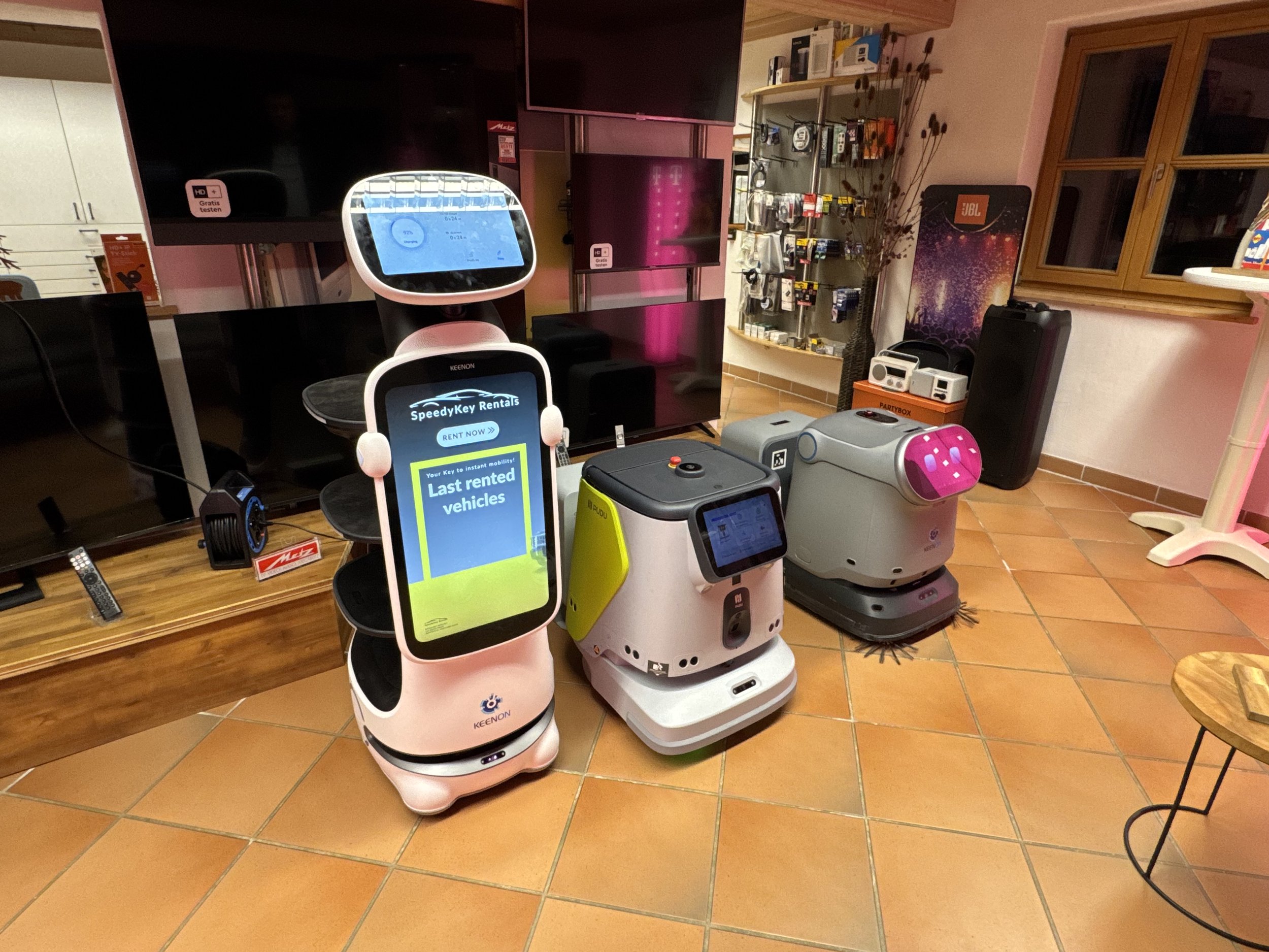 Elektrische Haushaltsgeräte und Roboter in einem Laden, darunter ein Roomba Staubsauger und ein KI-basierter Assistent auf einem Bildschirm.