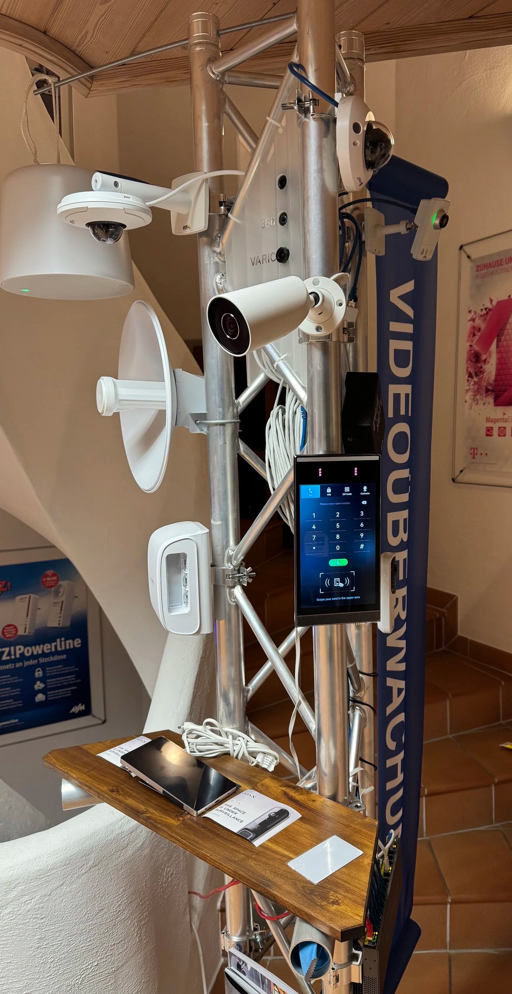Das Bild zeigt eine technische Ausrüstung mit mehreren Kameras, einem Touchscreen-Display, Kabeln und einem Holz-Tisch, auf dem ein Smartphone, eine Broschüre und ein kleiner weiße Gegenstand liegen. Es scheint sich um eine Präsentation oder Demonstration technischer Geräte zu handeln.