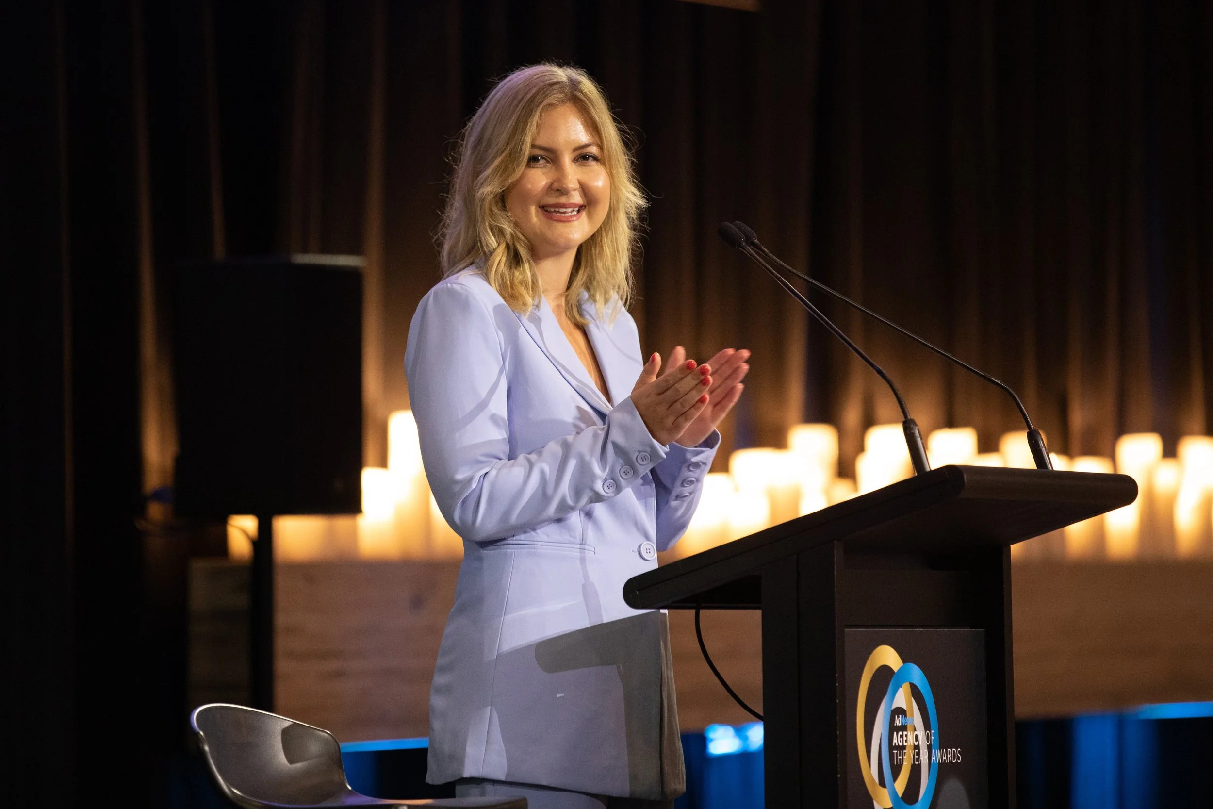 A woman with blonde hair wearing a light blue blazer speaking at a podium with microphones on a stage.
