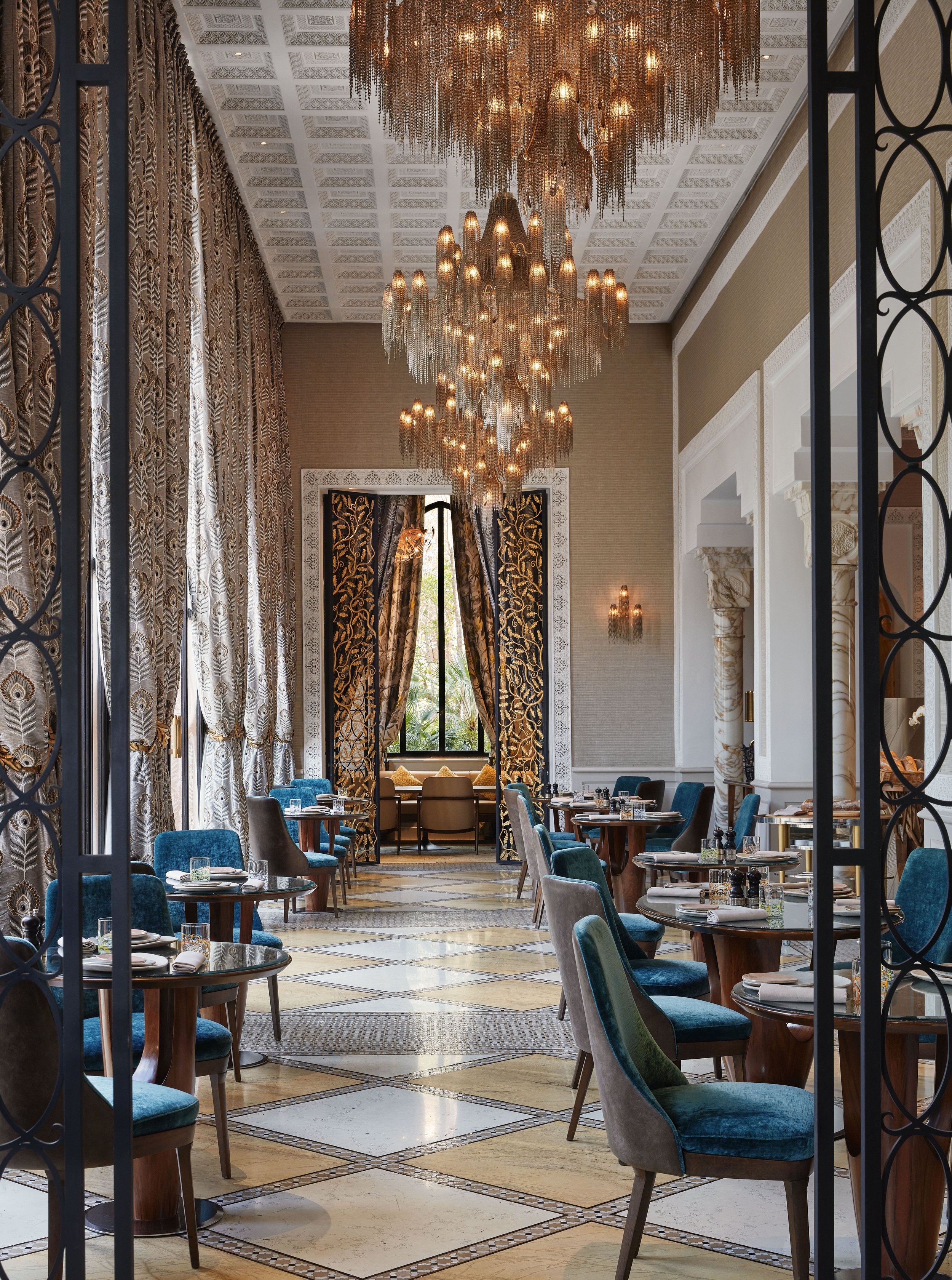 Elegant restaurant interior with a large crystal chandelier, ornate curtains, and neatly arranged tables with teal and gray chairs.