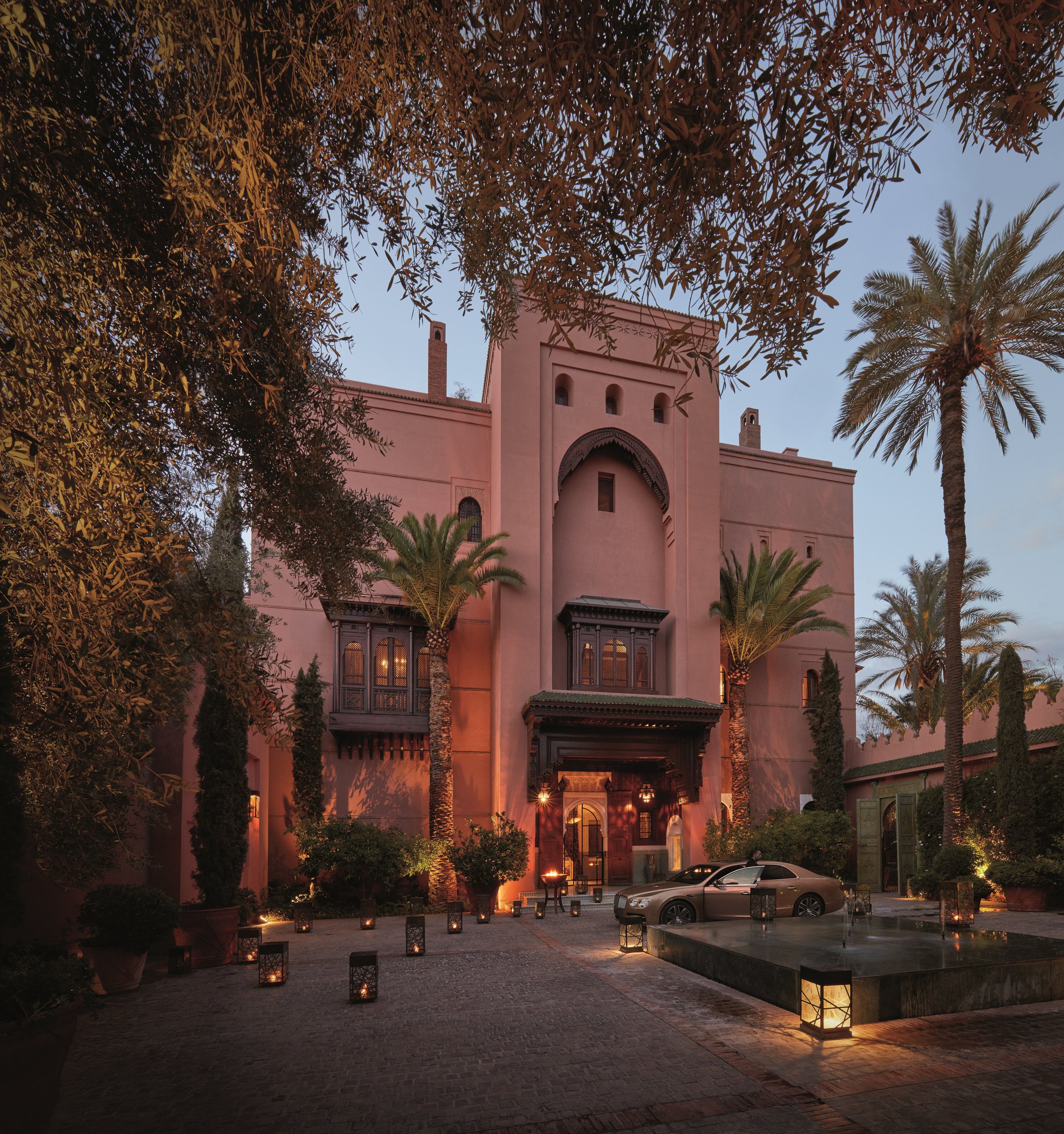 An evening view of a pink building with Moroccan architectural features, surrounded by palm trees and trees, illuminated by courtyard lighting, with lanterns lining a path leading to the building.