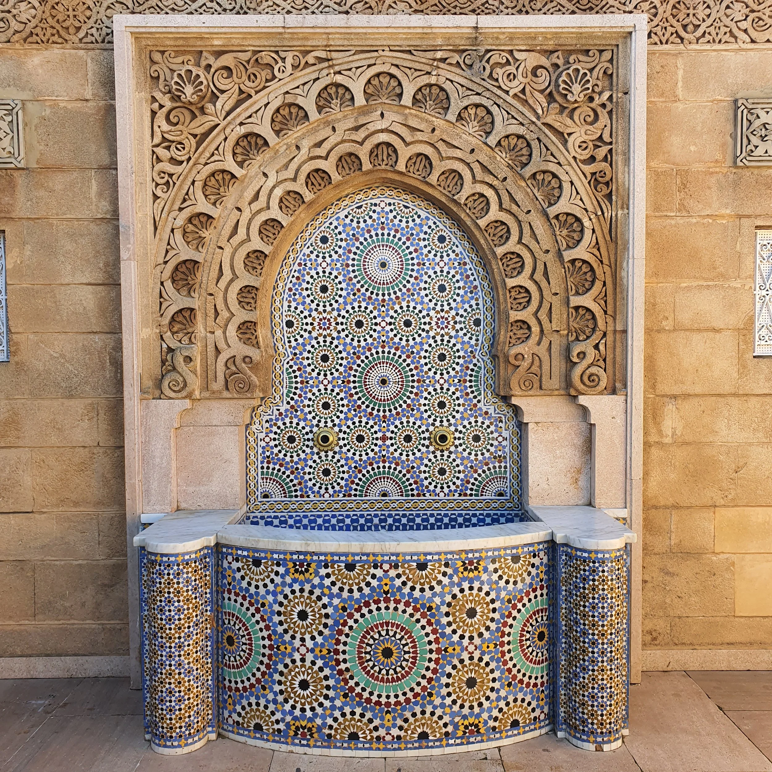 Intricate stone fountain with colorful mosaic tile design in geometric pattern, framed by carved stone arch.