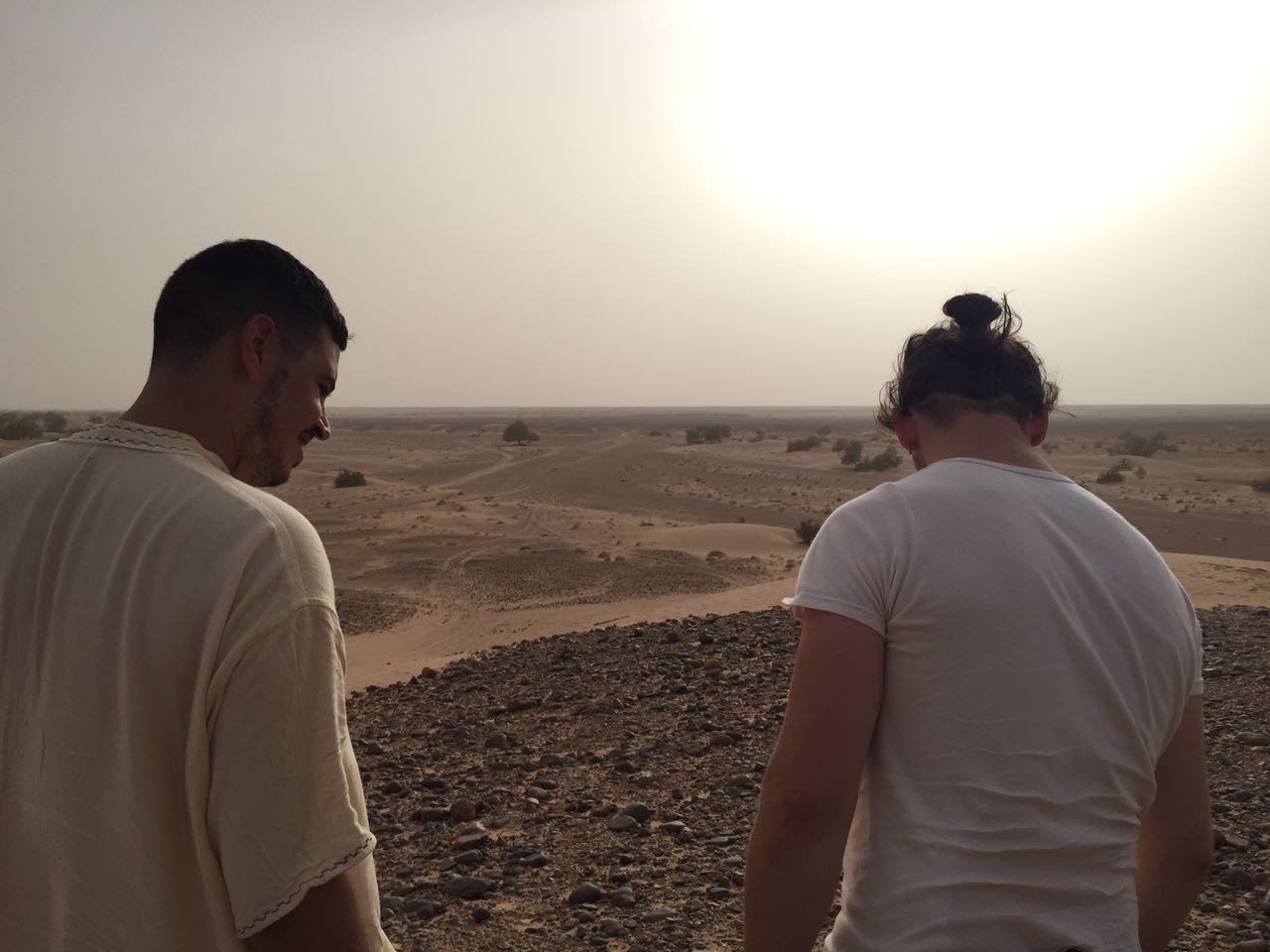 Two people standing on a rocky hill overlooking a desert landscape with sparse vegetation and a few trees in the distance.