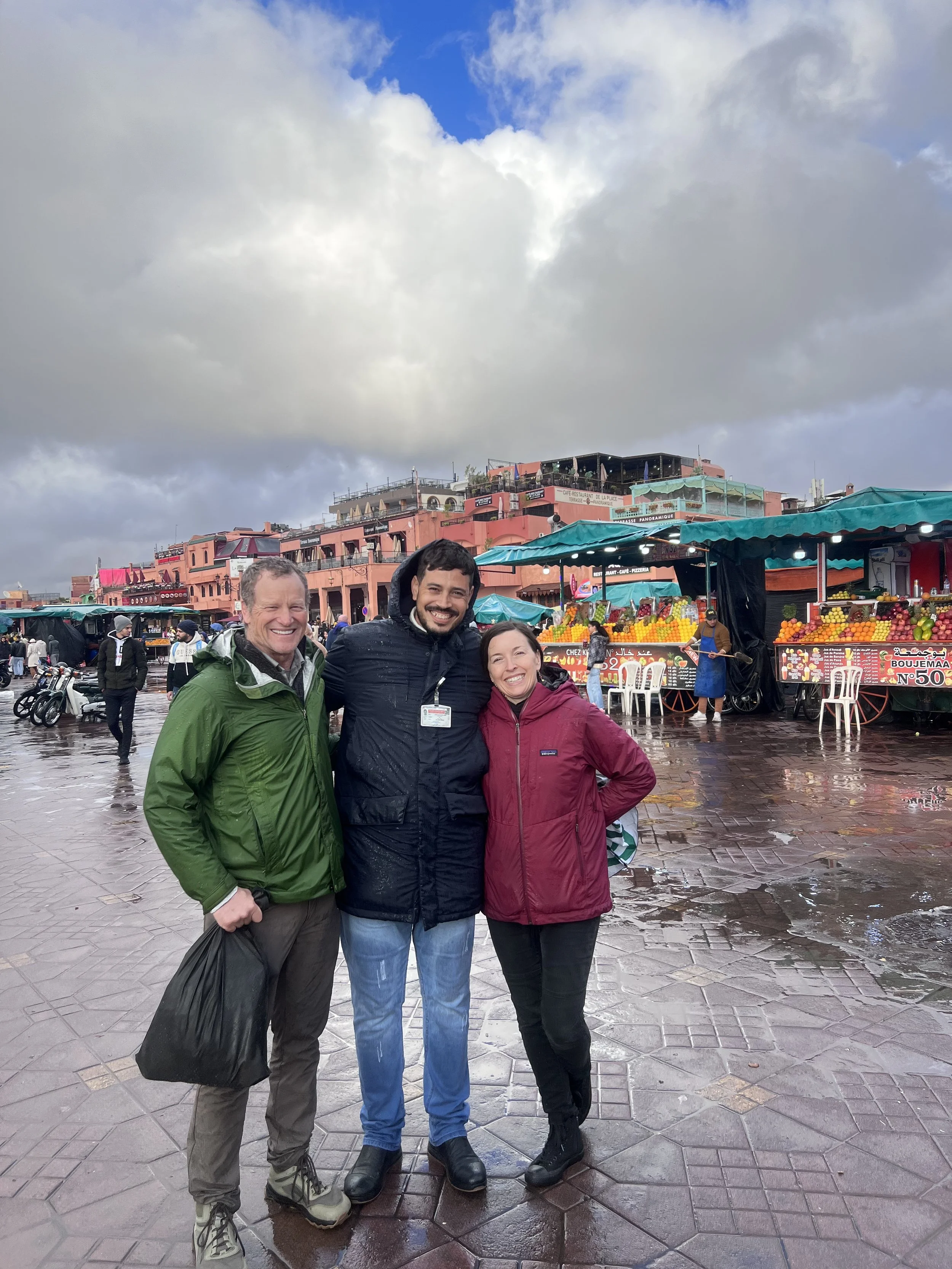Three people standing together outdoors in a public market area, smiling, with stalls selling fruit behind them and a cloudy sky overhead.