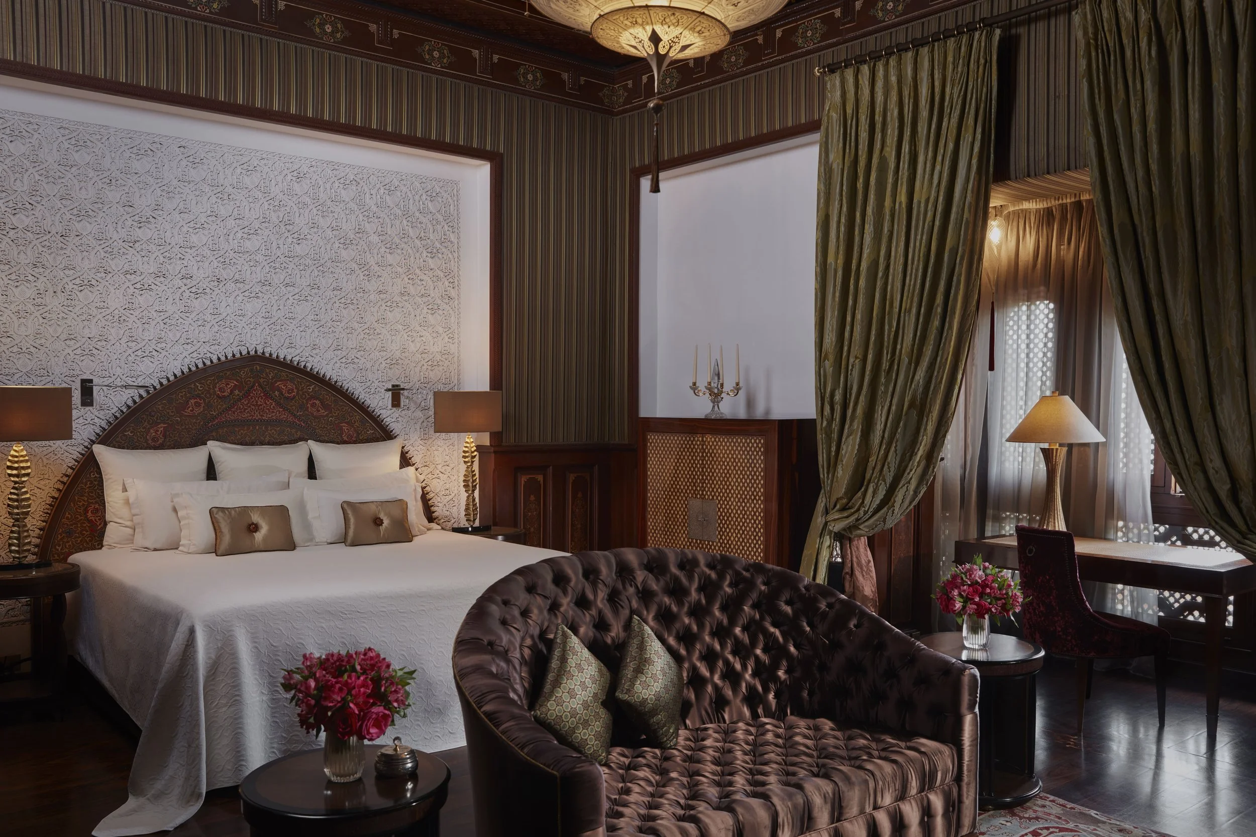 A luxurious hotel room features a large bed with white linens and decorative pillows, a tufted leather armchair with patterned cushions, side tables with lamps, a floral arrangement on a table, textured wall panels, striped and floral curtains, and o