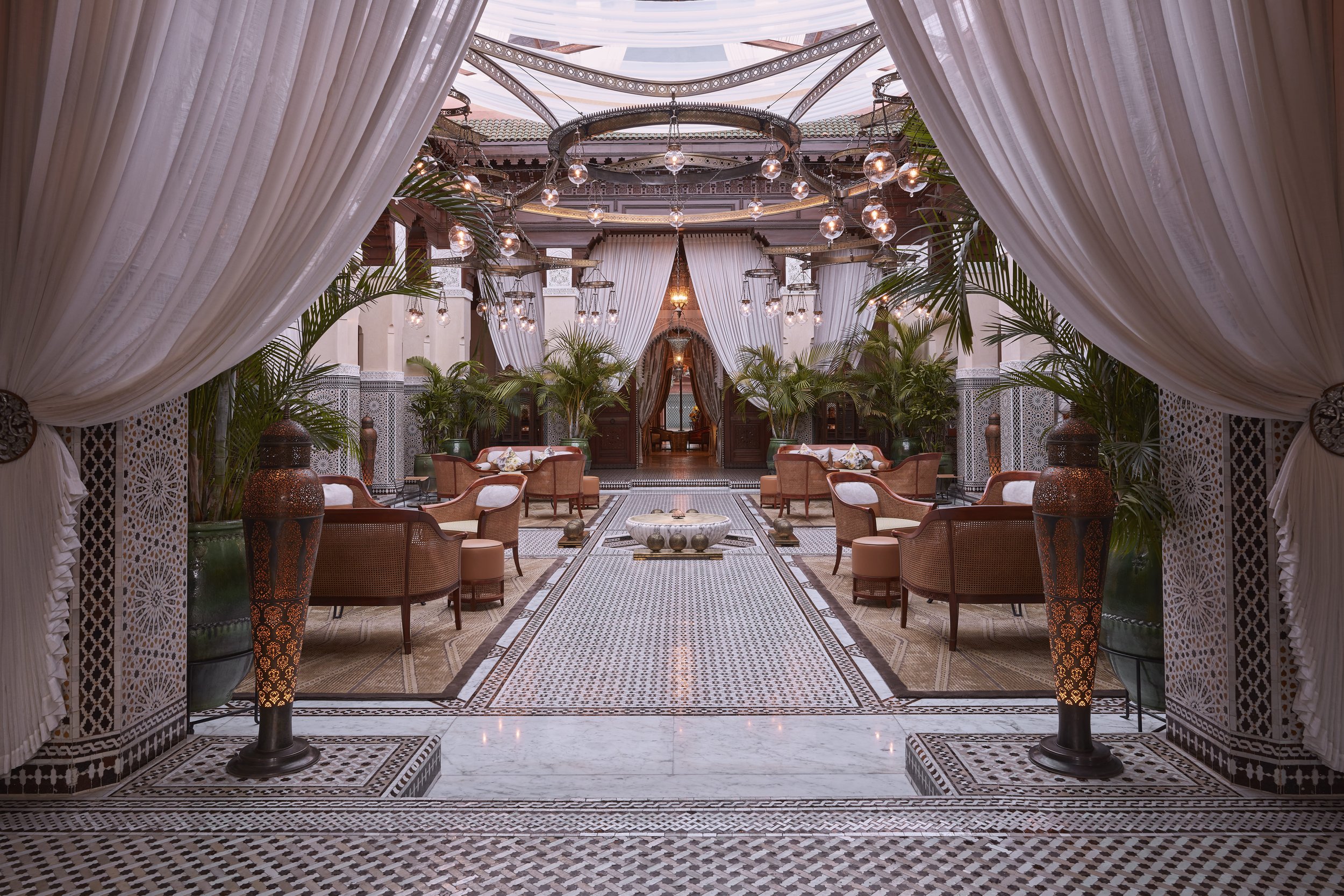 Elegant indoor lounge area with Moroccan-inspired design, featuring ornate tile flooring, lush green plants, white drapes, and circular chandeliers hanging from a decorative ceiling.
