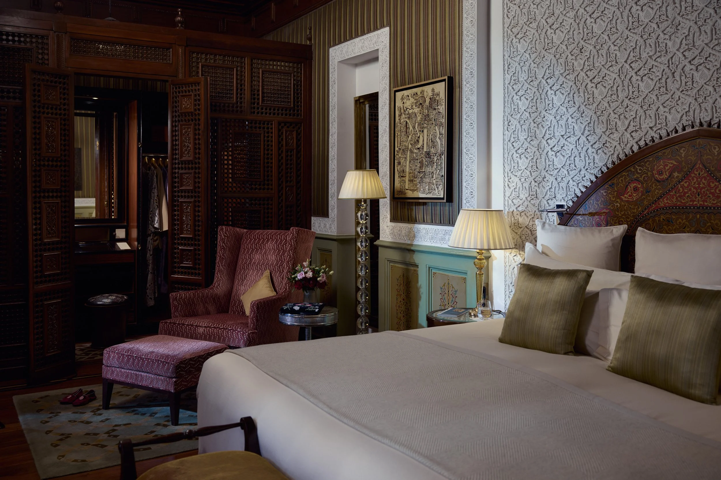 A luxurious bedroom with ornate decor, a large bed with multiple pillows, two table lamps, a red upholstered armchair, a floral centerpiece, patterned wallpaper, framed art, and a wooden wardrobe with intricate lattice doors.