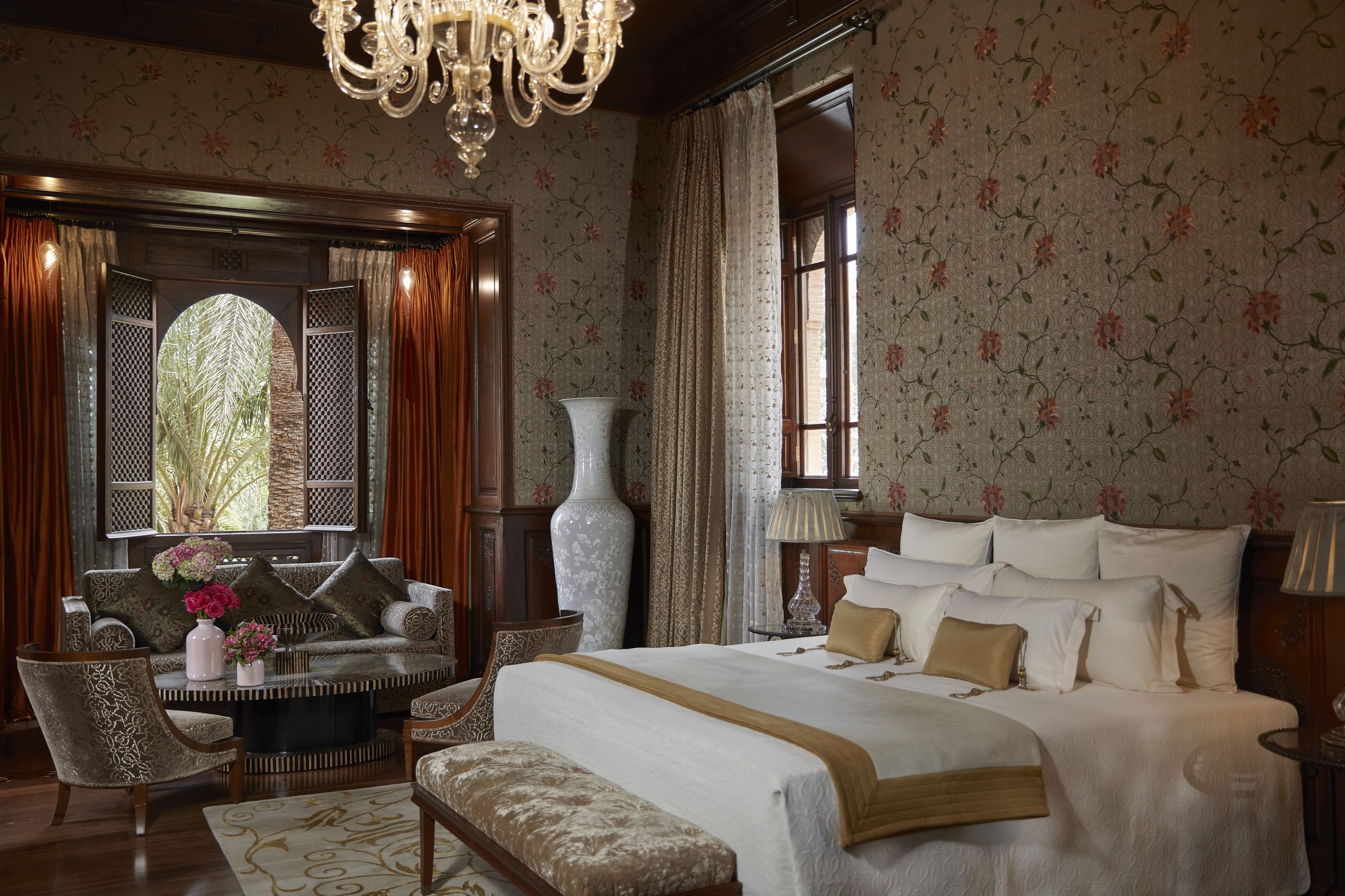 Elegant living room with floral wallpaper, chandelier, wooden framed windows with curtains, white sofa with pillows, a small table with a lamp, and a seating area with floral arrangements.