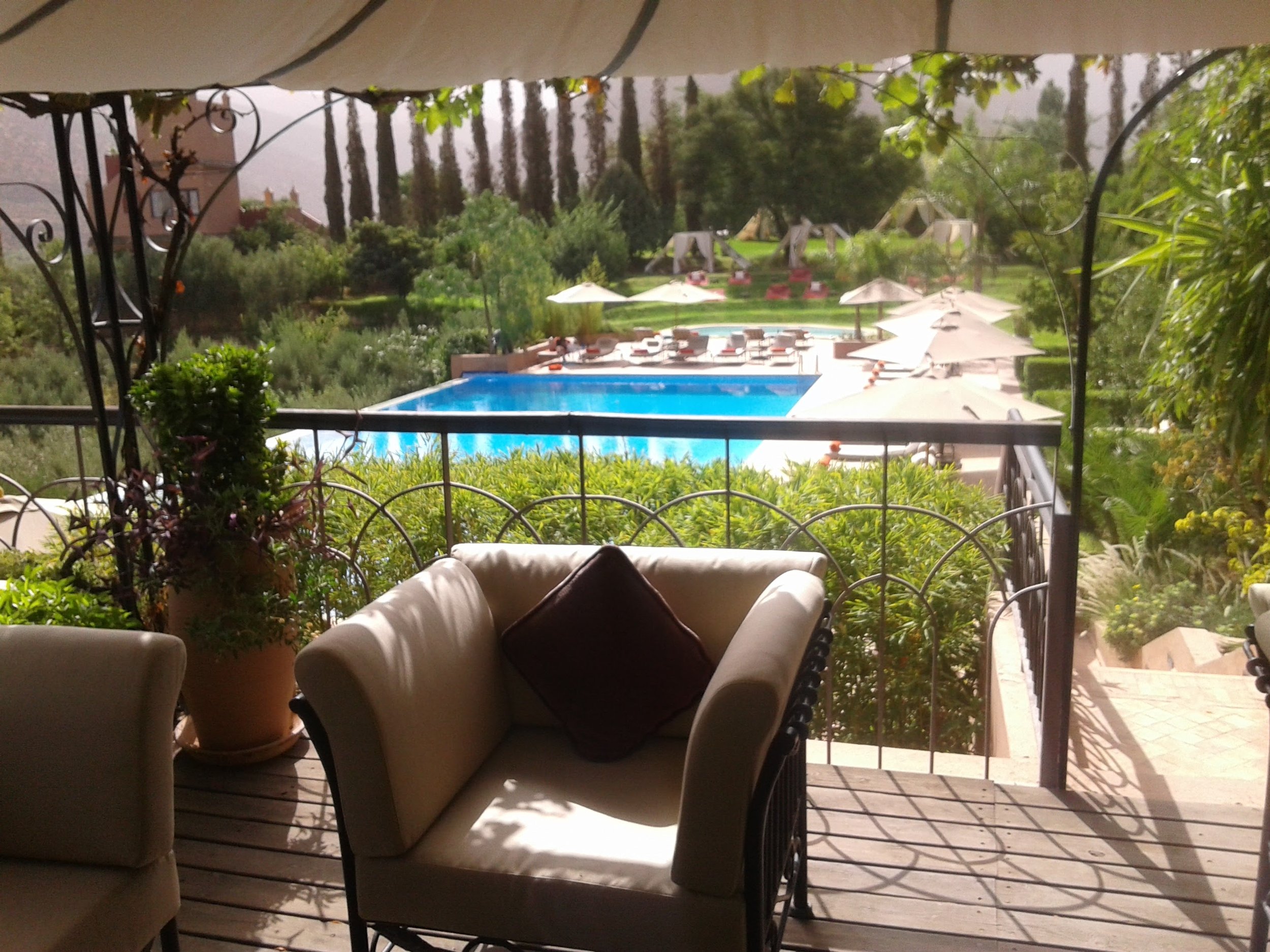 Interior view from a covered patio or balcony overlooking a swimming pool, outdoor furniture, umbrellas, lush greenery, tall trees, and a distant building.