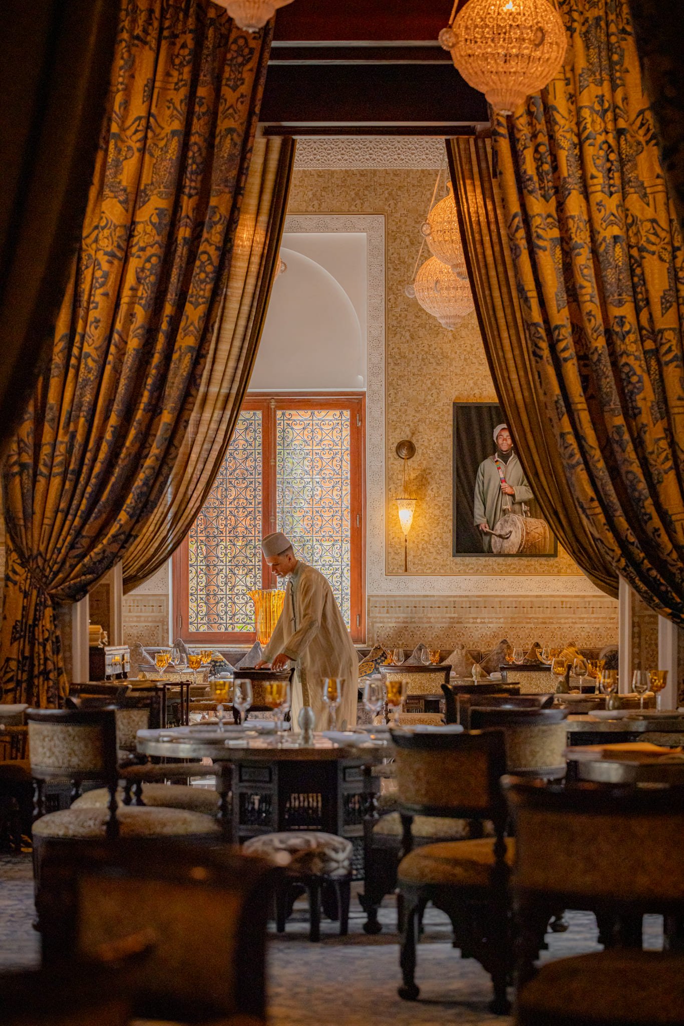 Interior view of an elegant restaurant with ornate curtains, chandeliers, and a large portrait of a man playing a traditional drum on the wall. A waiter in traditional attire is setting the table, which is decorated with wine glasses and tableware.
