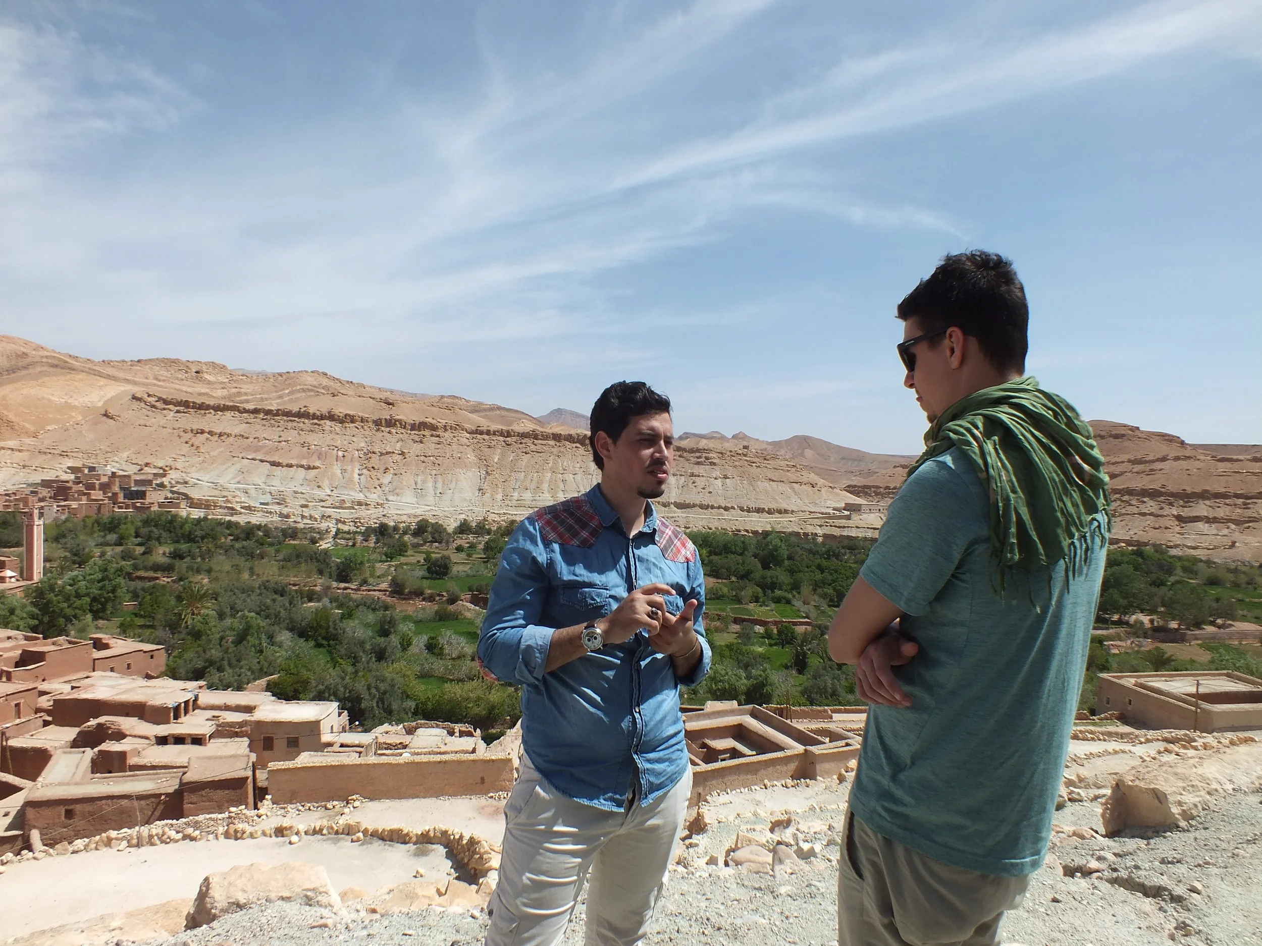 Two men stand outdoors in a desert landscape with traditional mud buildings and rocky hills in the background. One man faces the camera holding a phone, while the other stands with his arms crossed wearing sunglasses and a scarf.