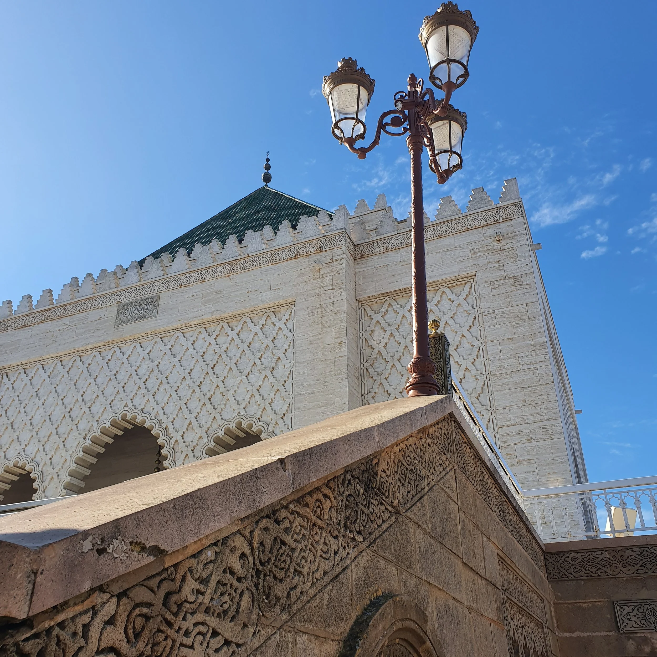 Close-up of ornate white stone building with intricate carvings, a green tiled roof, and a decorative streetlamp against a blue sky.
