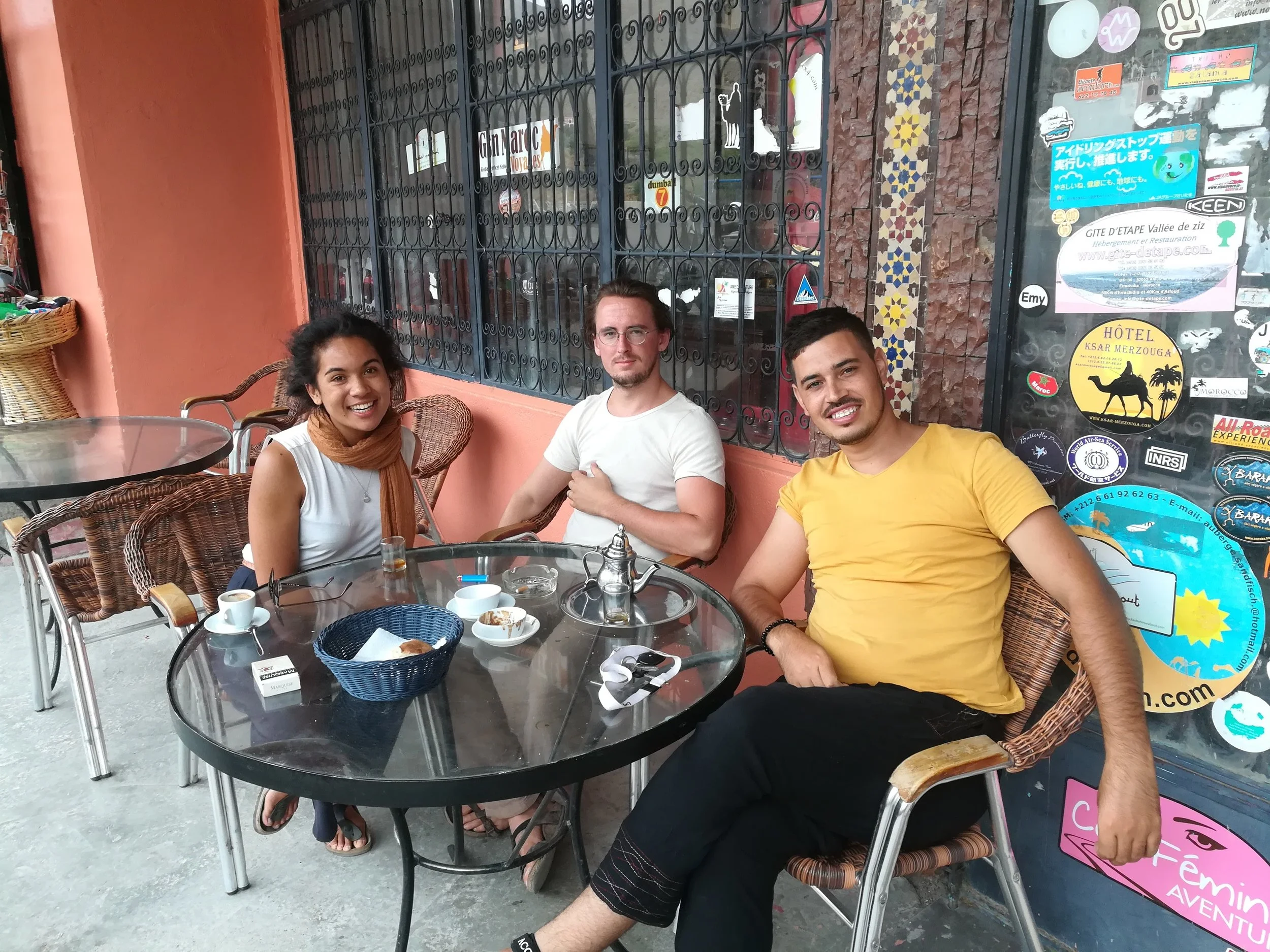 Three young people sitting at a round glass table outside a building with a decorative tile wall and a window covered with stickers. The group appears to be enjoying a casual meal or coffee, with cups and small plates on the table.