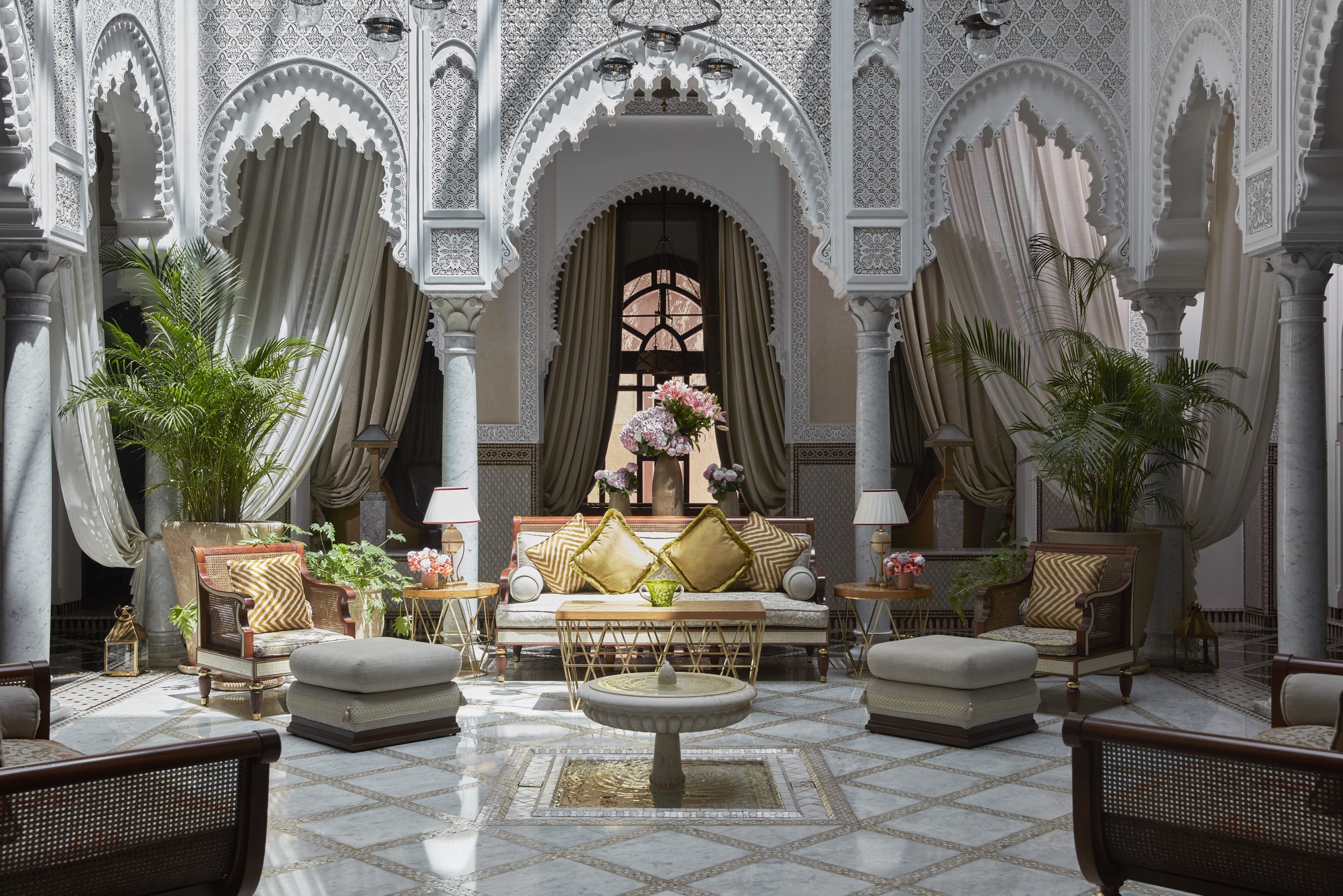 Elegant indoor sitting area with white ornate arches, draped curtains, and lush green plants. There is a central table with a flower arrangement and multiple comfortable chairs and sofas with yellow and gold cushions.