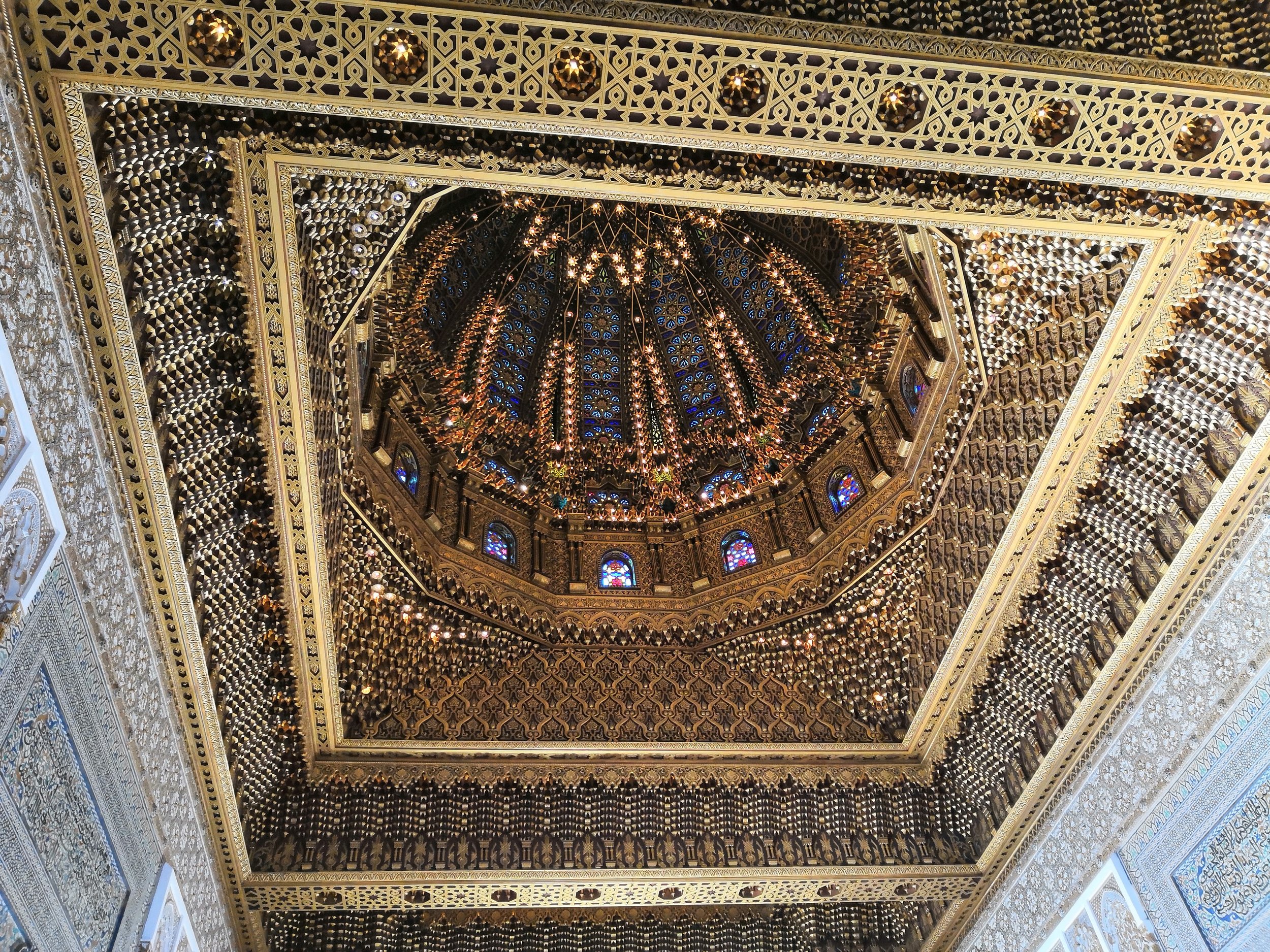 Intricate golden ceiling with geometric patterns, stained glass windows, and hanging lights, viewed from below.