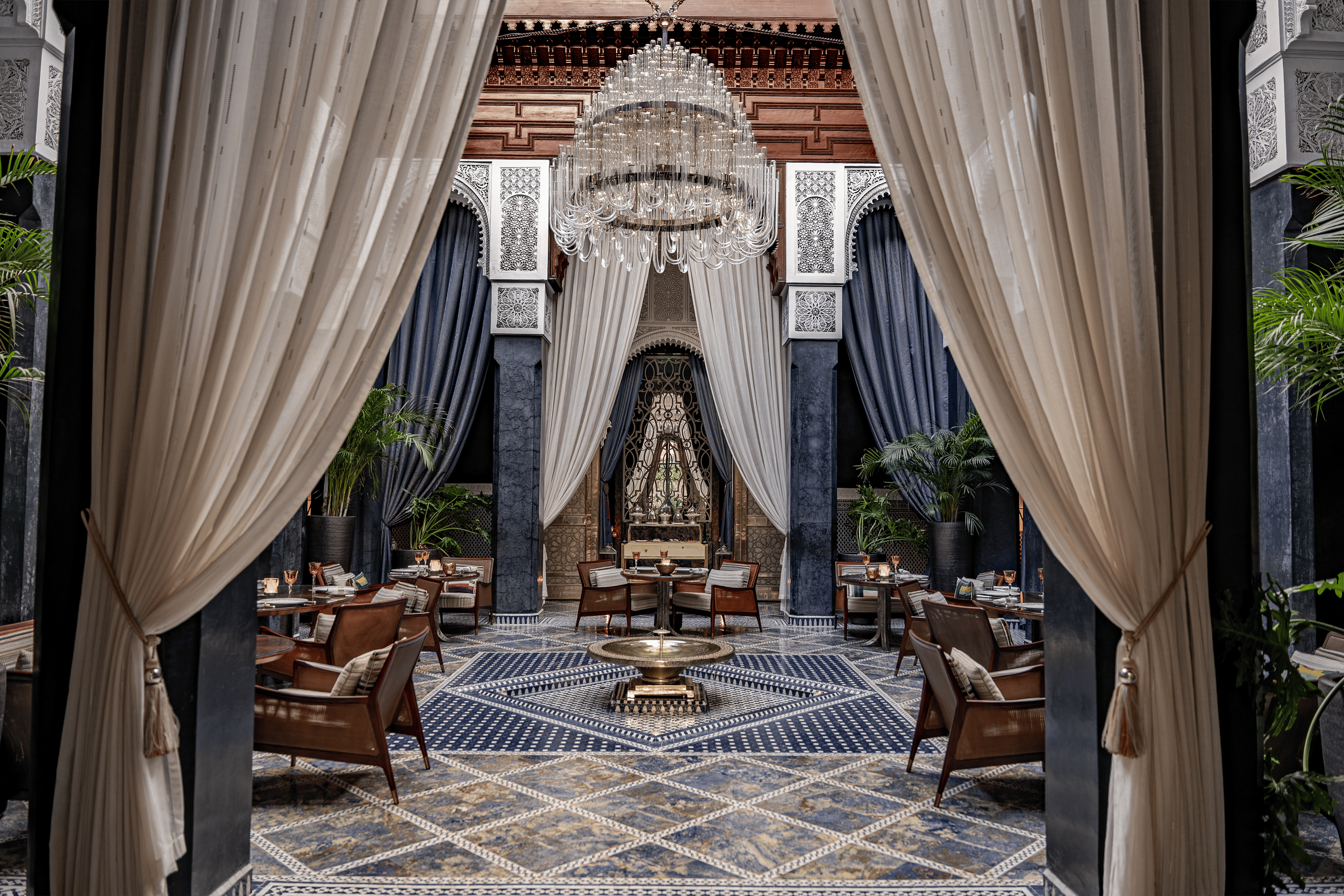 Elegant restaurant interior with a grand chandelier, draped curtains, adorned arches, and a patterned tile floor.