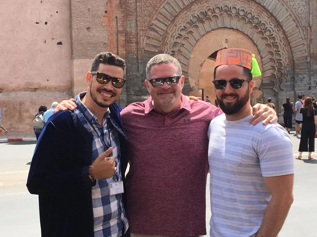 Three men standing close together outdoors, smiling, with their arms around each other's shoulders. The background features a large stone arch and a person wearing a colorful hat.