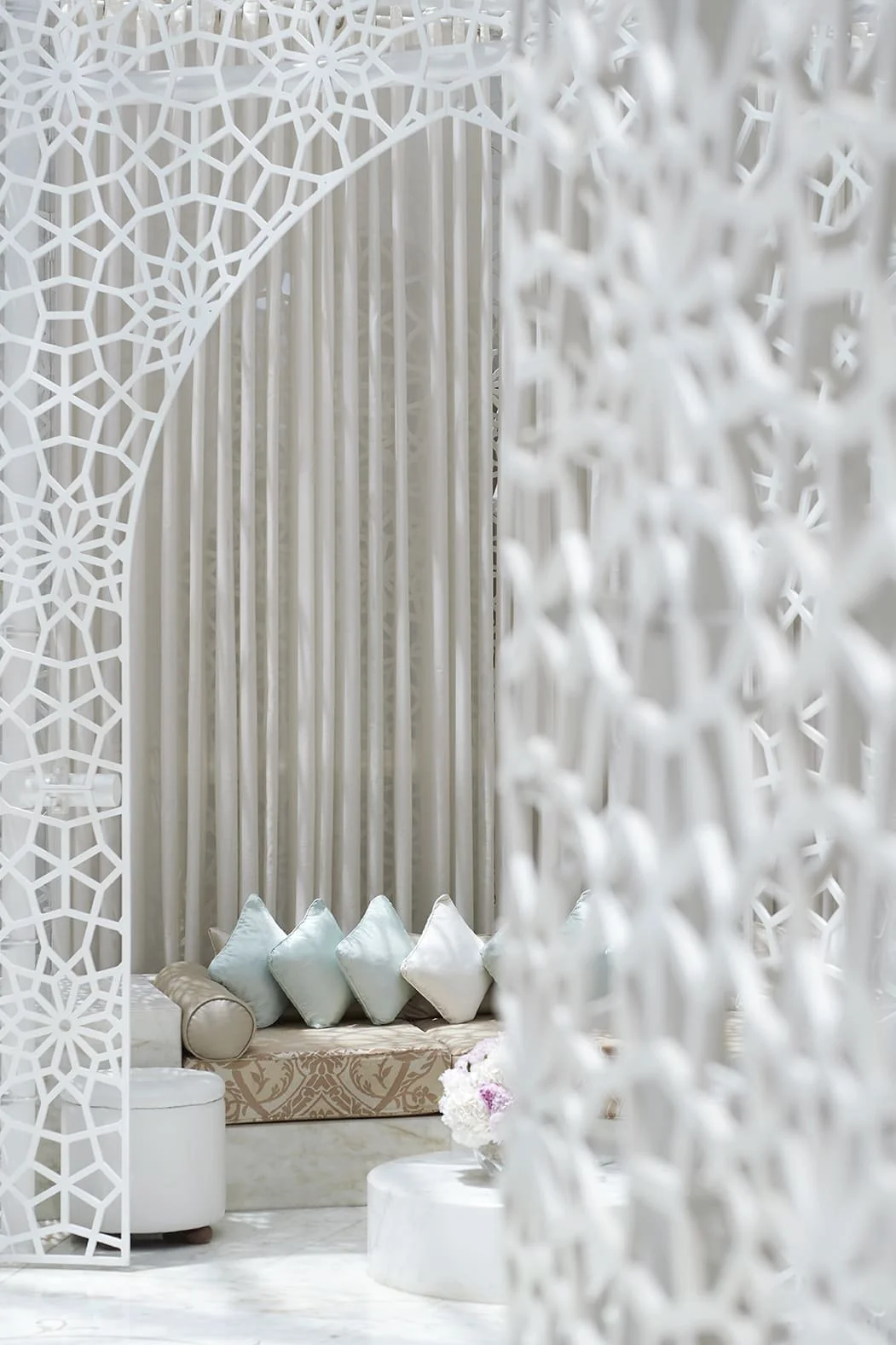 Elegant indoor seating area with white ornate lattice walls, a beige patterned bench with pastel pillows, and a round marble table with a flower arrangement.