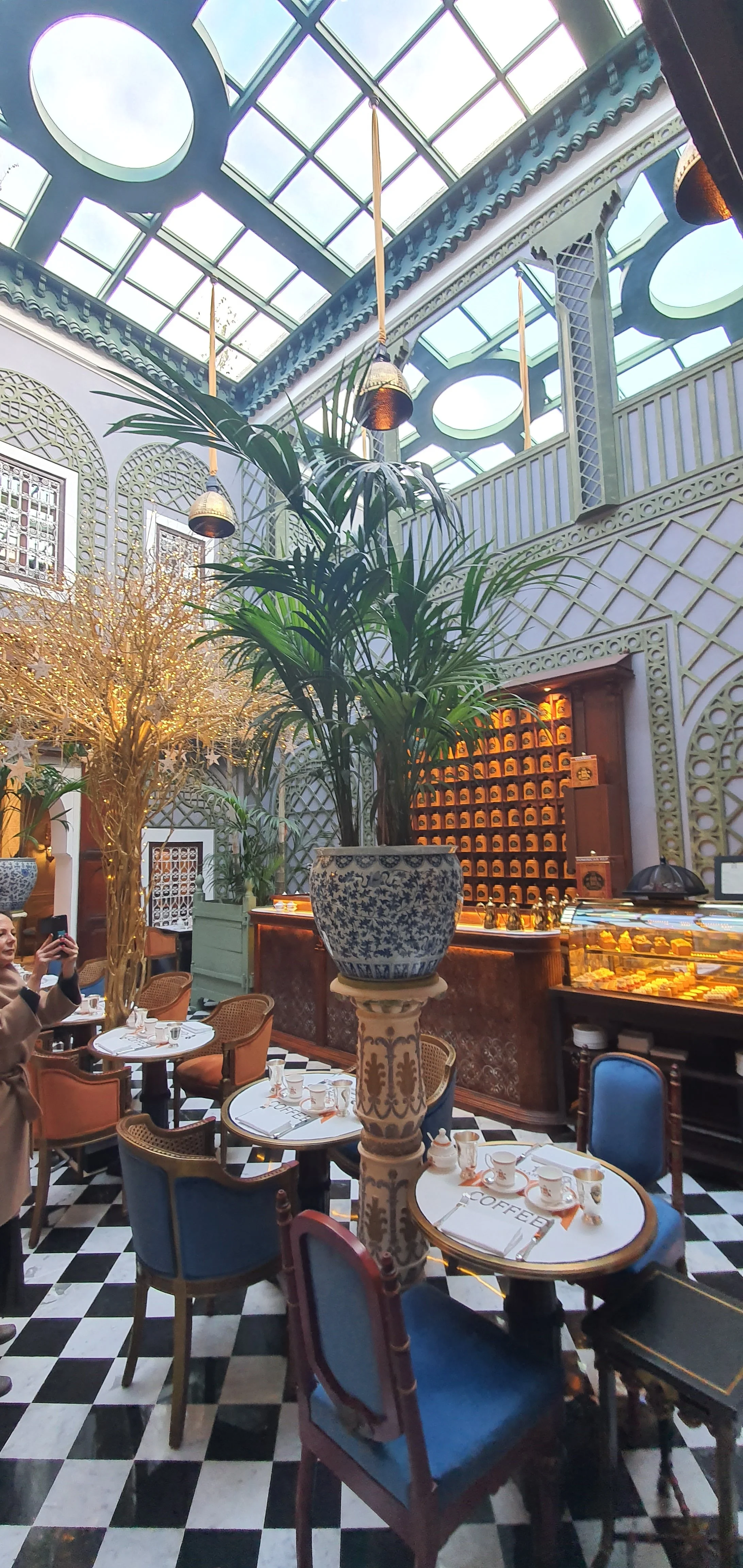 Interior of a restaurant or cafe with black and white checkered floor, large potted plants, decorative walls, a glass ceiling allowing natural light, tables set with cups and dishes, and a woman taking a photo.