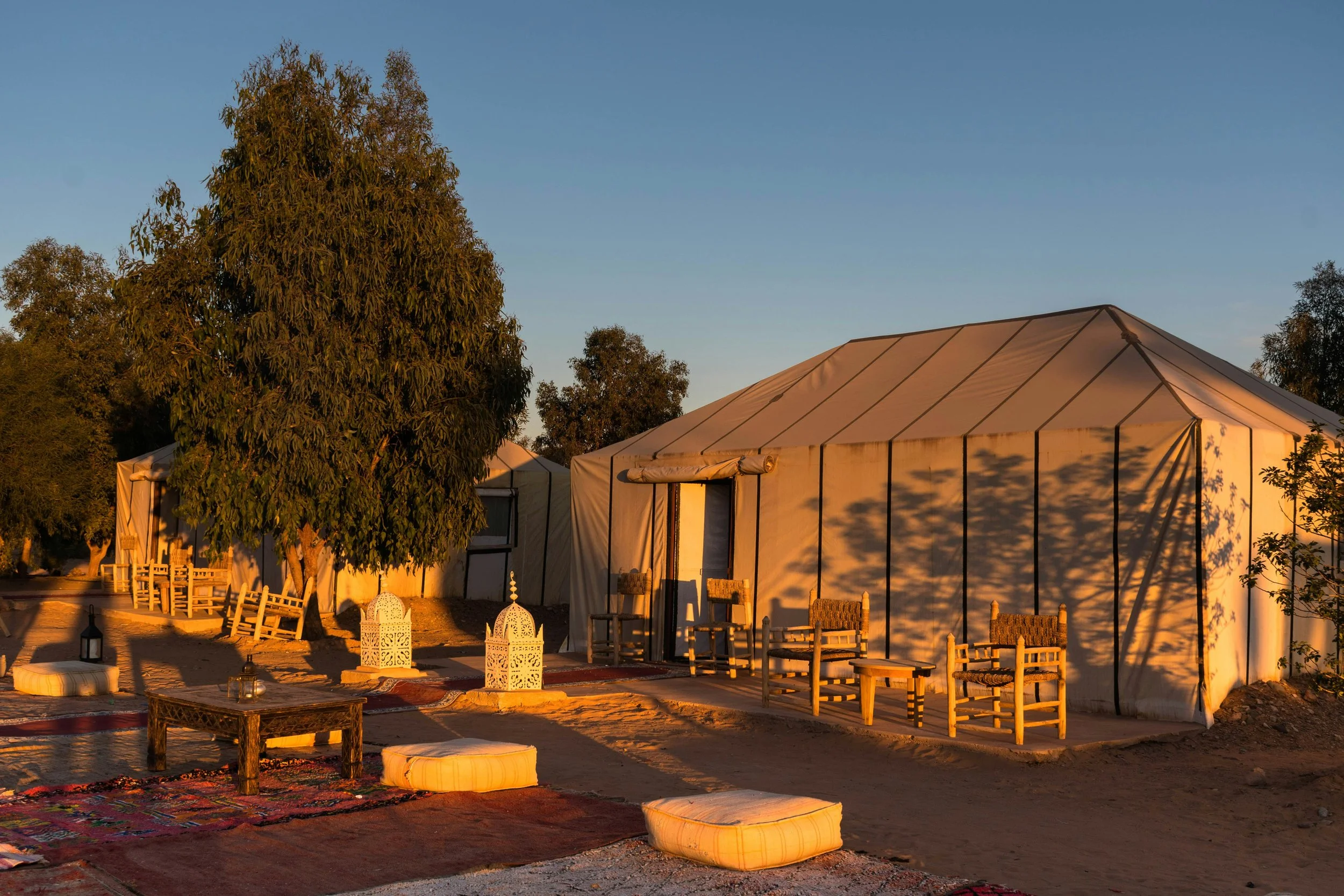 An outdoor setting during sunset with a large white tent, surrounded by trees and chairs, illuminated by warm sunlight, and decorated with lanterns and rugs.