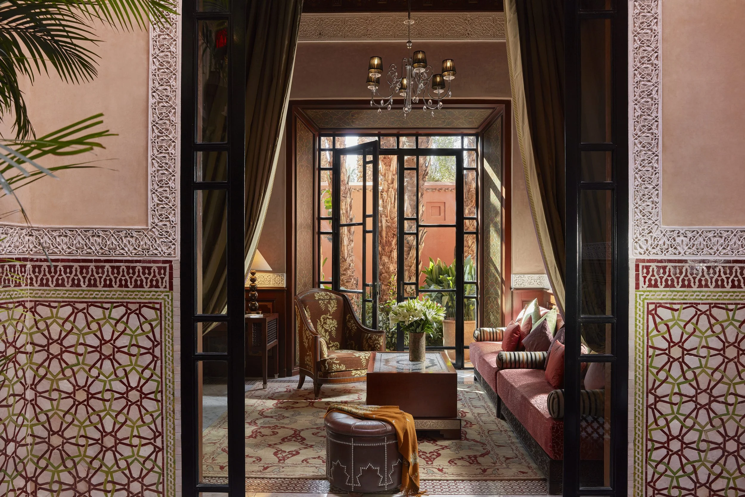 Interior of a luxurious room with large glass doors opening to an outdoor area with trees. Inside, there are elegant furniture pieces including armchairs, a sofa, a lamp, a coffee table with a flower arrangement, and decorative carpets. A chandelier 